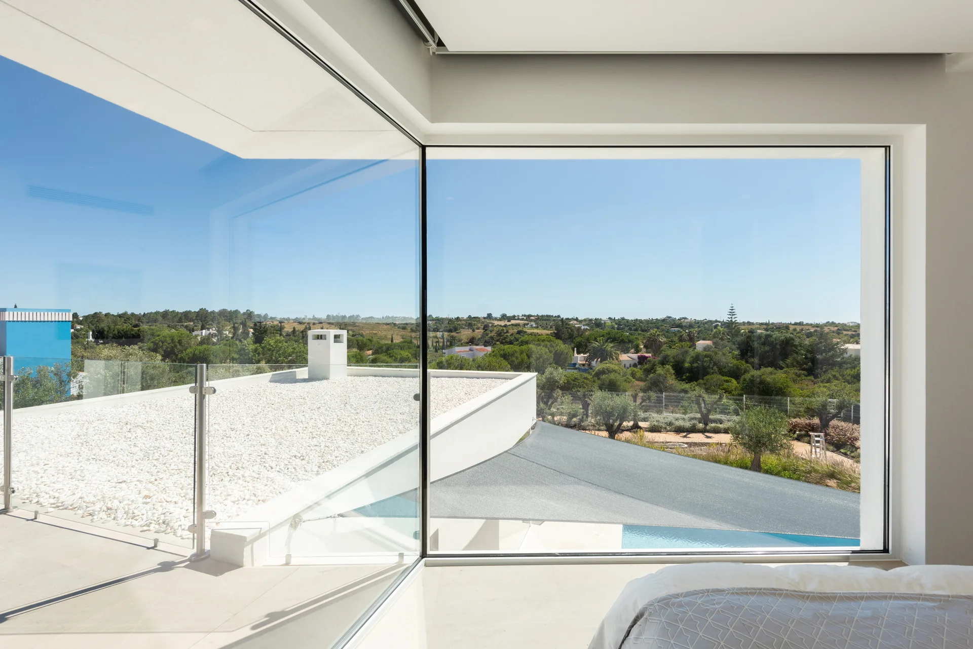 A modern bedroom features a floor-to-ceiling corner glass window looking out over a white gravel roof terrace and a green landscape.
