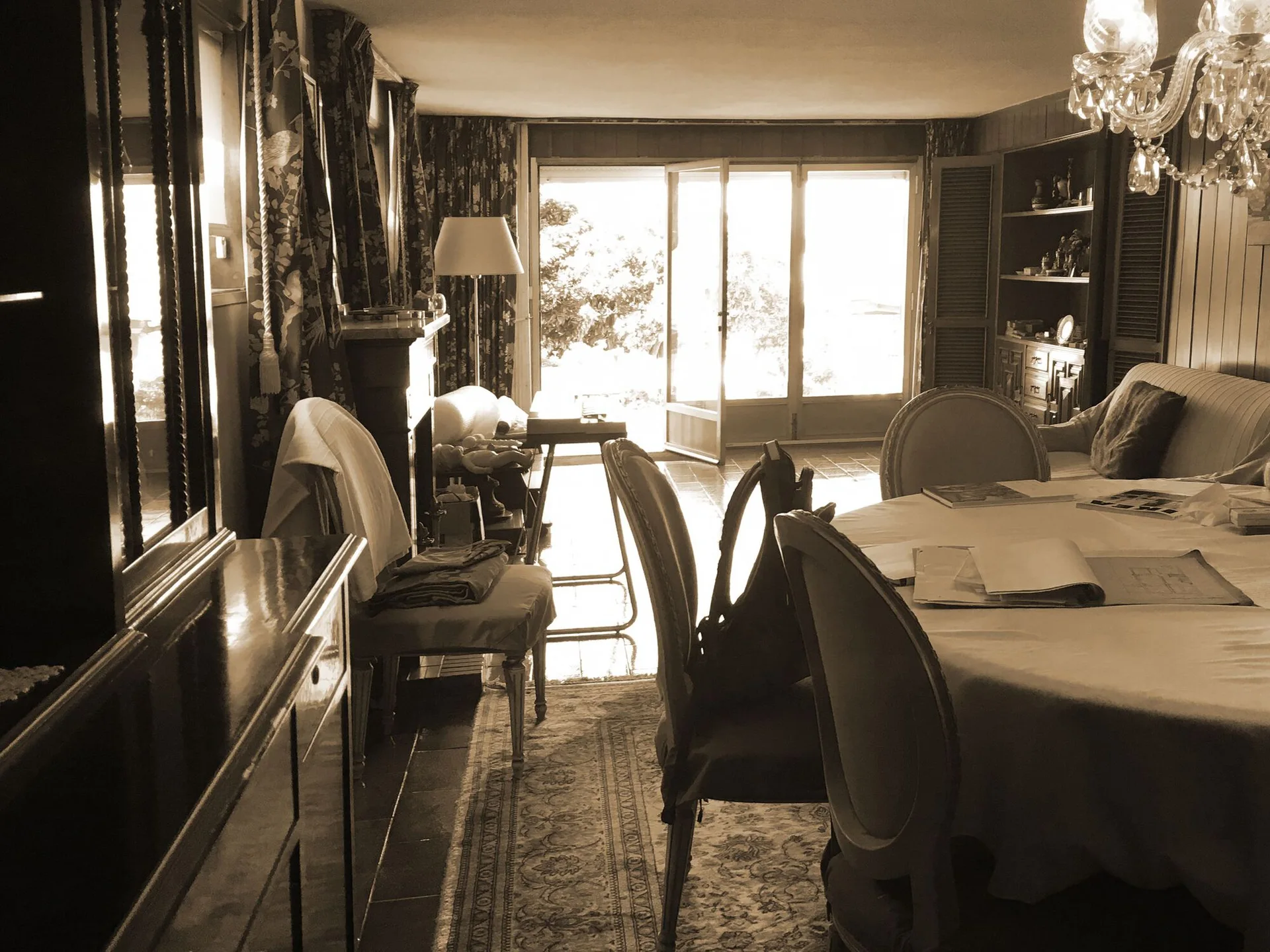 A sepia-toned traditional dining room features a long table with a white cloth, a crystal chandelier, wood-paneled walls, and open glass French doors.