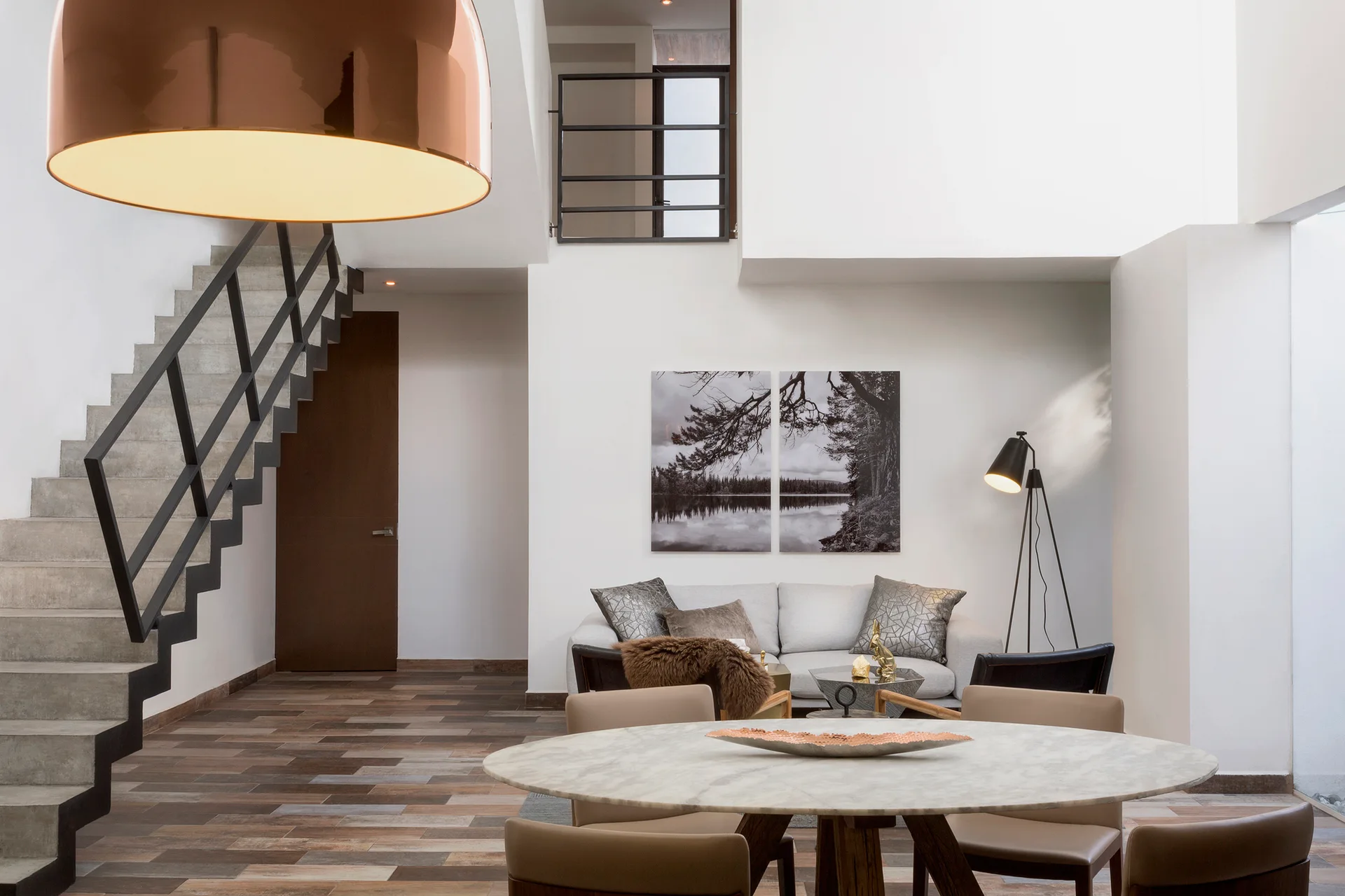 An open-plan living and dining space featuring a concrete staircase, a round marble-like table, a gray sofa, and a large copper pendant light.