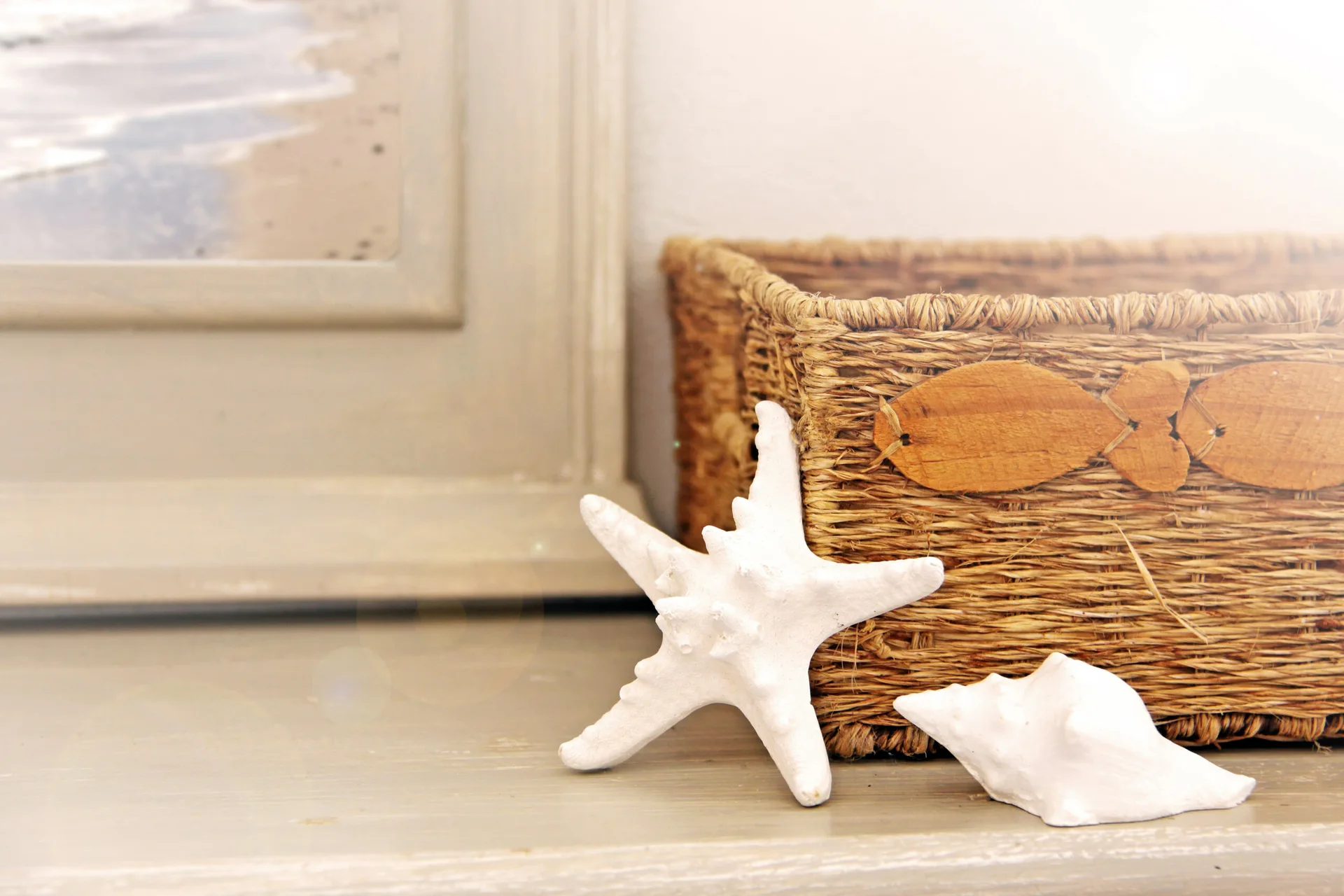 A white starfish and seashell rest next to a woven wicker basket on a light-colored wooden shelf beside a framed picture.