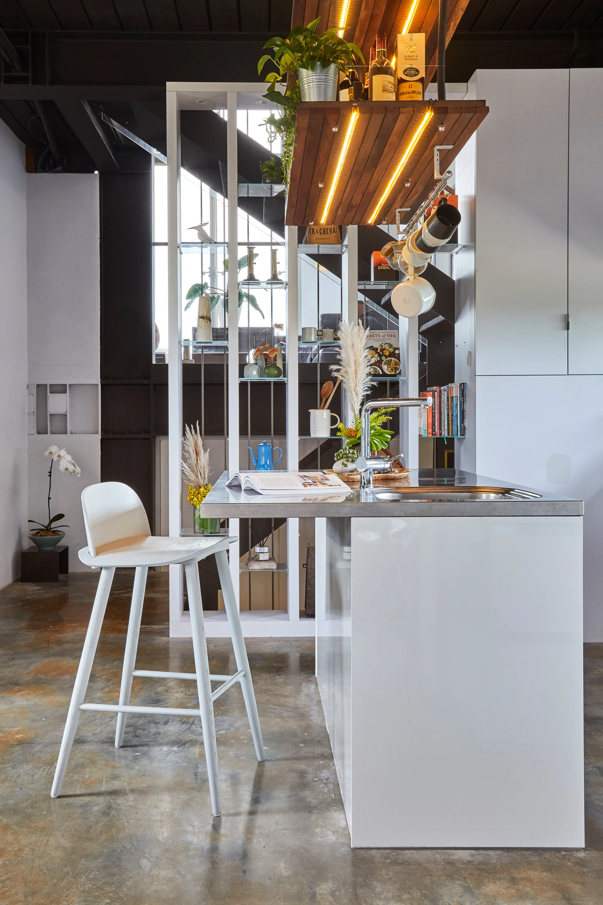 A modern kitchen featuring a white island with a stainless steel sink, a white wooden bar stool, and an open wood shelving unit hanging overhead.