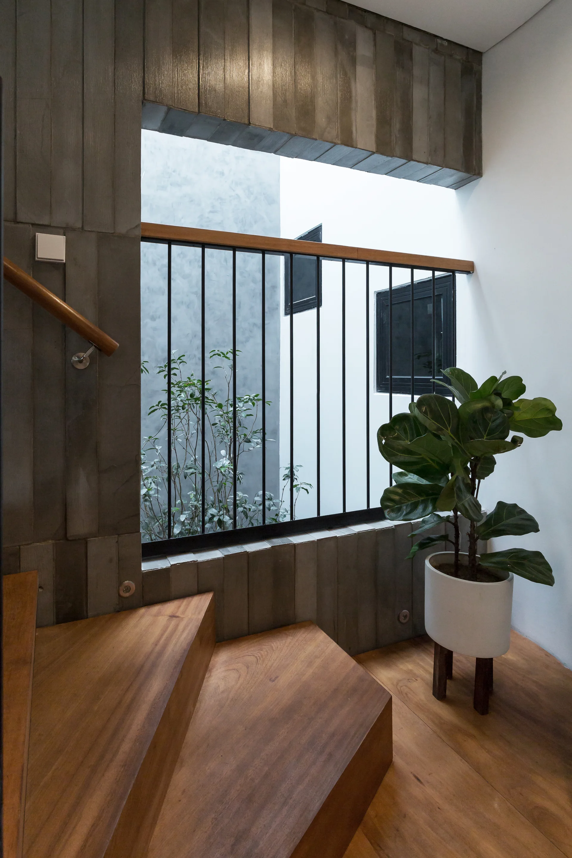 A modern staircase with wooden treads and a handrail set against a grey vertical-paneled wall next to a black metal railing and potted plant.