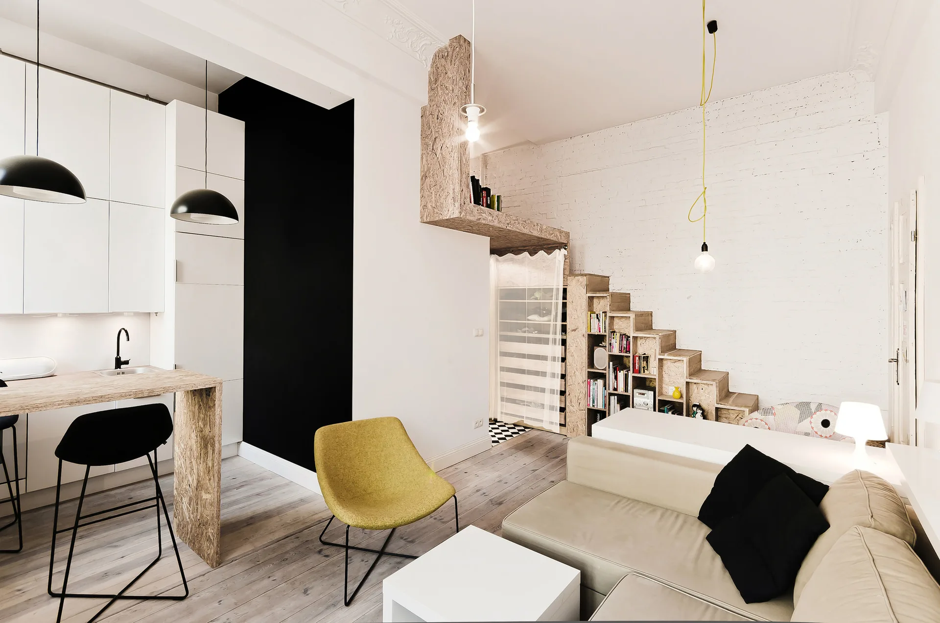 This studio apartment features a beige sofa, a yellow accent chair, and a custom OSB staircase with built-in bookshelves.