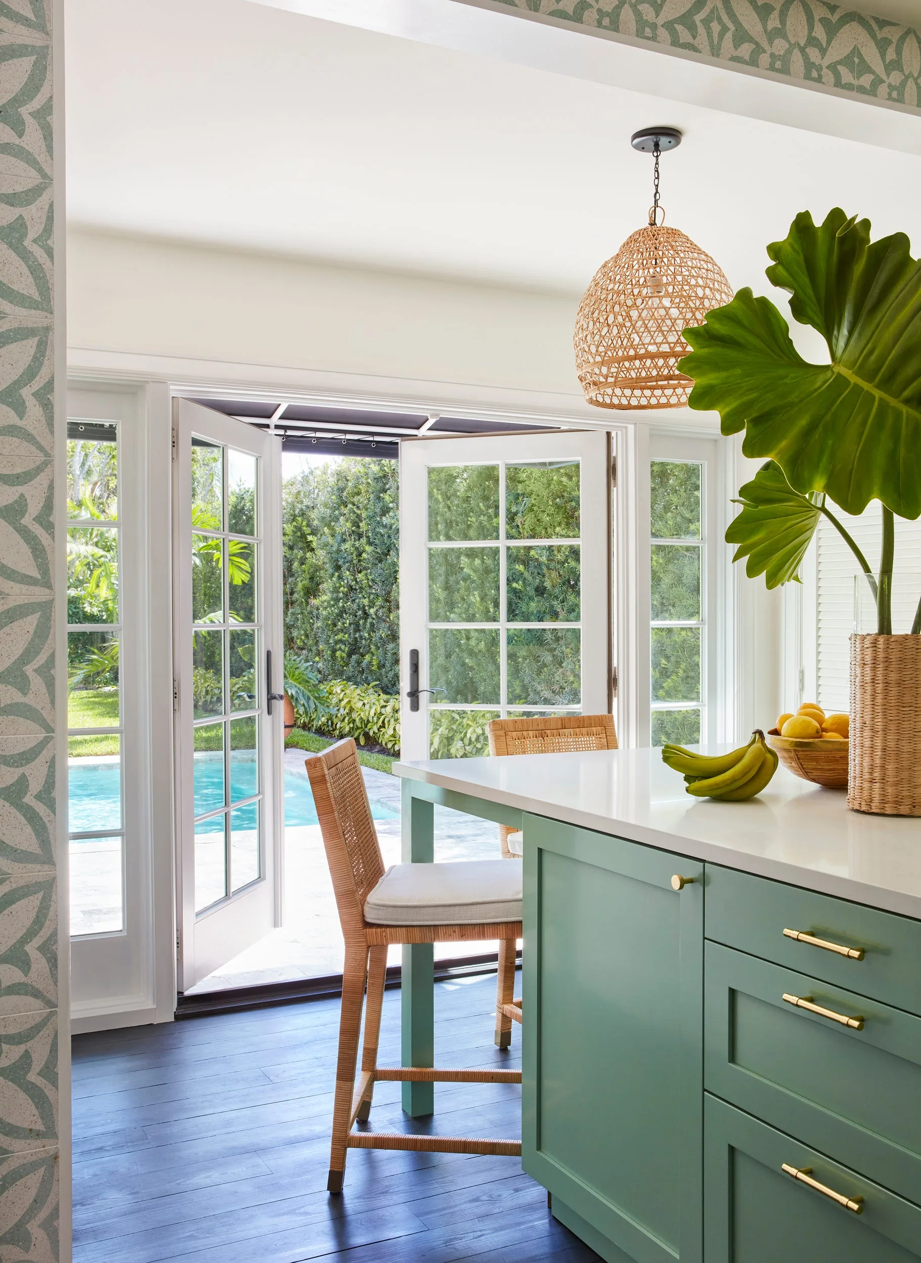 A bright kitchen features a green island with woven bar stools and open glass doors leading to an outdoor swimming pool.