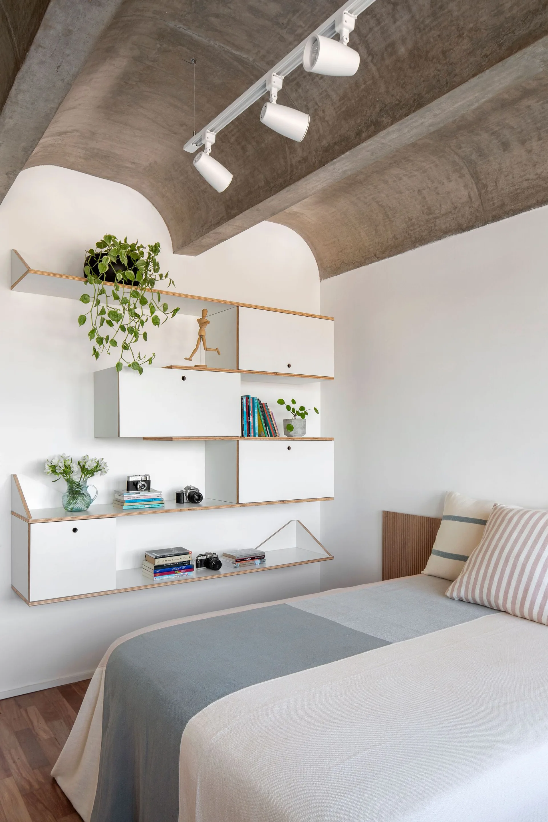 A minimalist bedroom features modular white plywood shelving on a white wall beneath a curved, vaulted concrete ceiling and a bed with striped pillows.