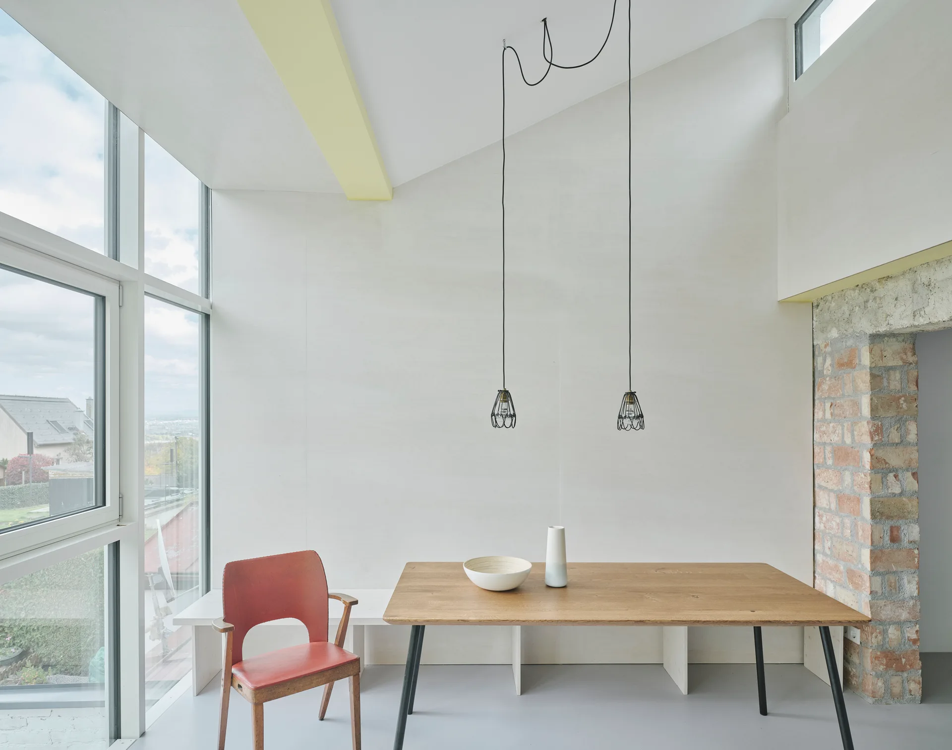 A minimalist dining room features a wooden table, a red mid-century chair, hanging cage pendant lights, and a large window.