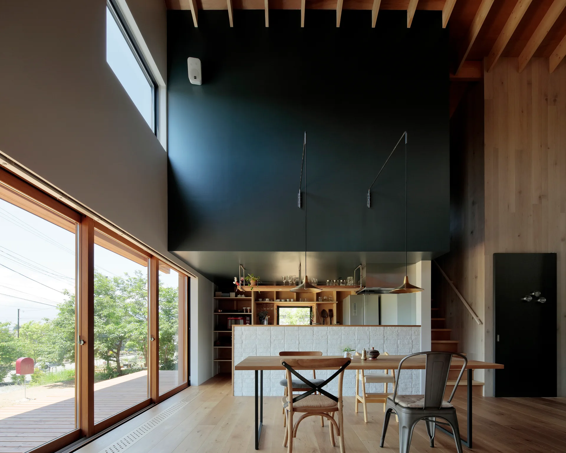 A modern dining room featuring a wood table, mismatched chairs, and a kitchen island beneath a large matte black architectural volume.