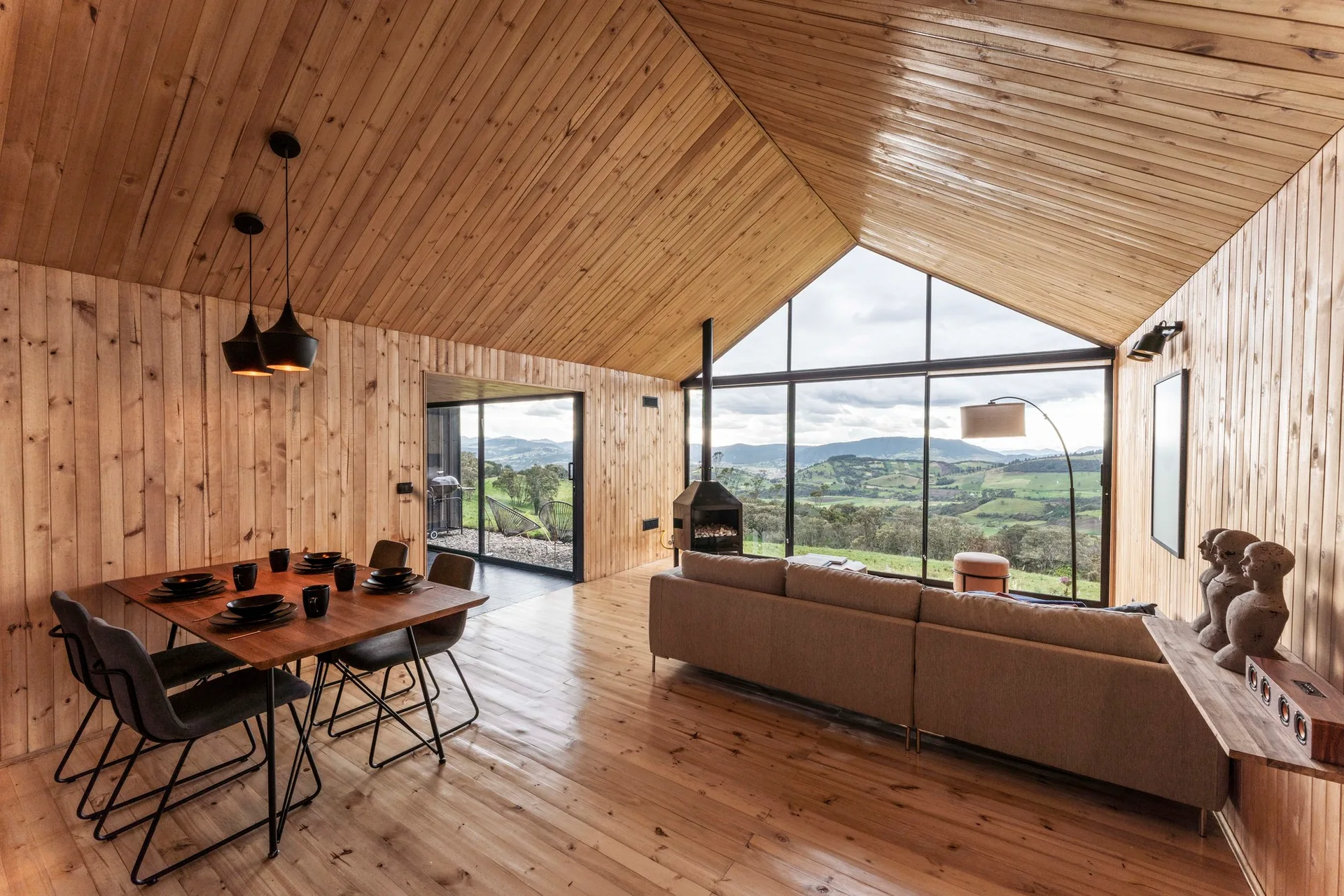 A wood-paneled living and dining area features a vaulted ceiling, a wooden dining table, and a beige sofa facing a large glass wall with hills outside.