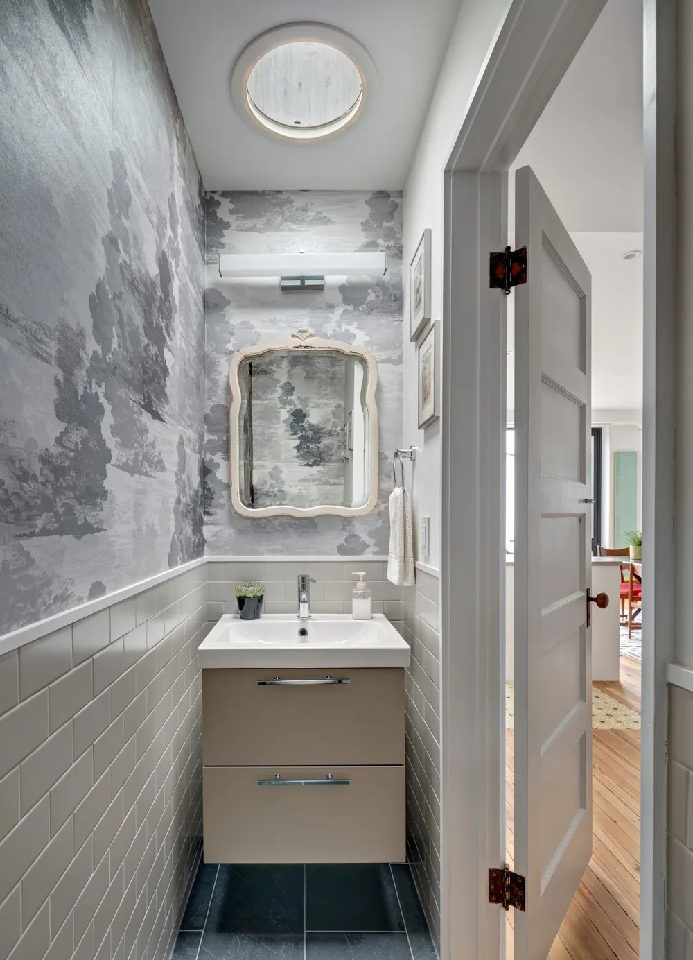 Narrow powder room with a beige wall-mounted vanity, gray landscape wallpaper, white subway tile wainscoting, and a circular skylight.