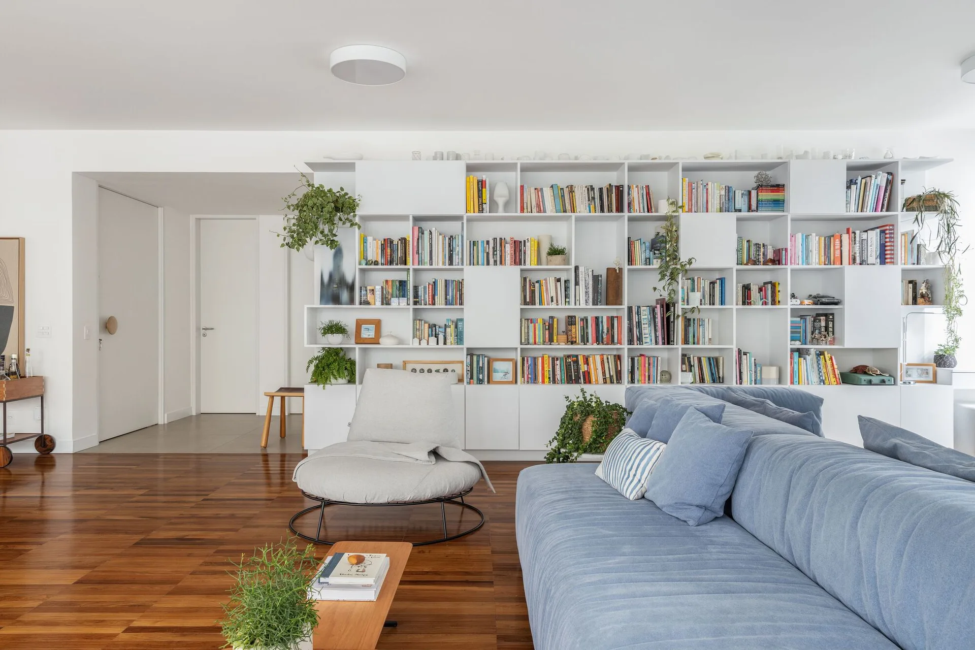 A contemporary living room features a blue sofa, a circular gray lounge chair, and a large white bookshelf on polished wood floors.