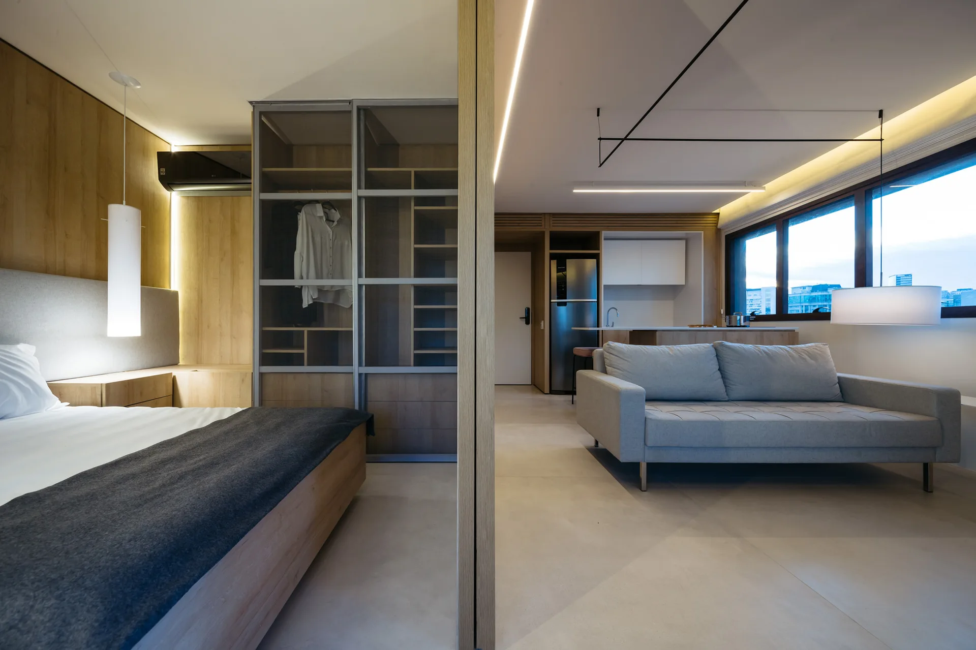 A modern studio apartment separated by a wooden sliding divider, featuring a bed, a glass-front wardrobe, and a grey sofa in the living area.