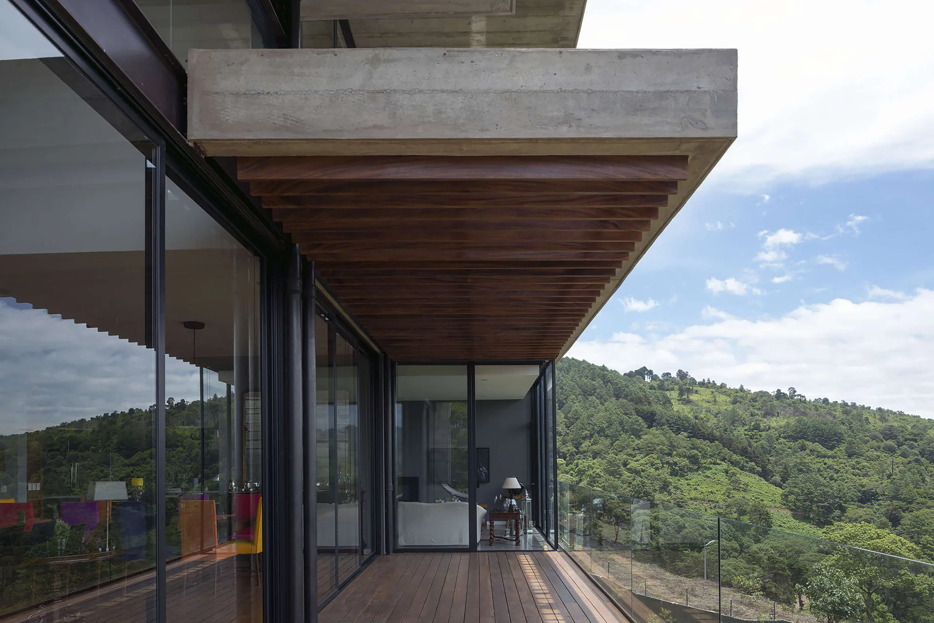 A modern outdoor terrace features a slatted wood ceiling, matching plank decking, and a glass railing overlooking a green mountain landscape.