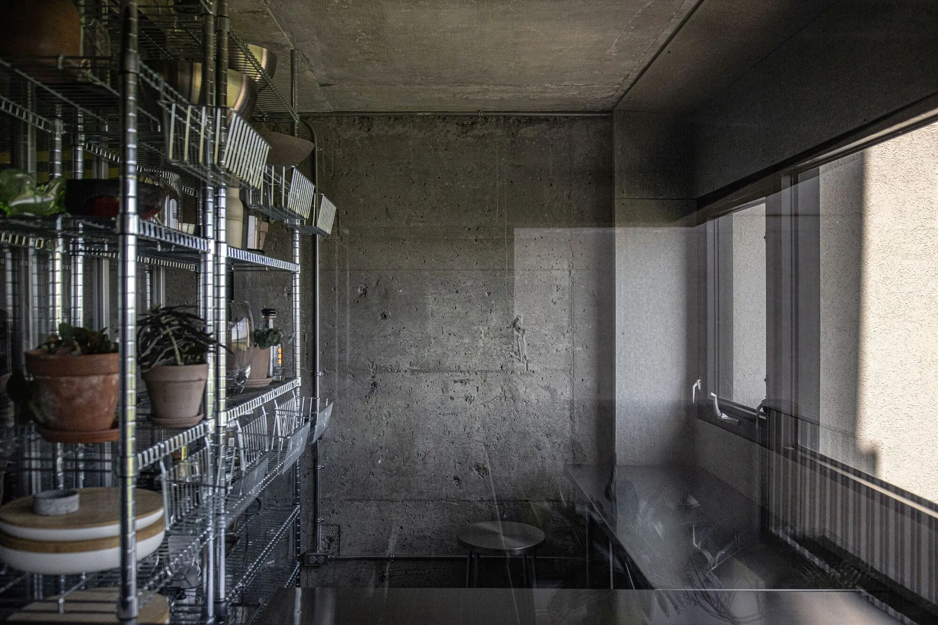 A minimalist industrial room features exposed raw concrete walls and a tall wire metal shelving unit holding terracotta plant pots and kitchenware.
