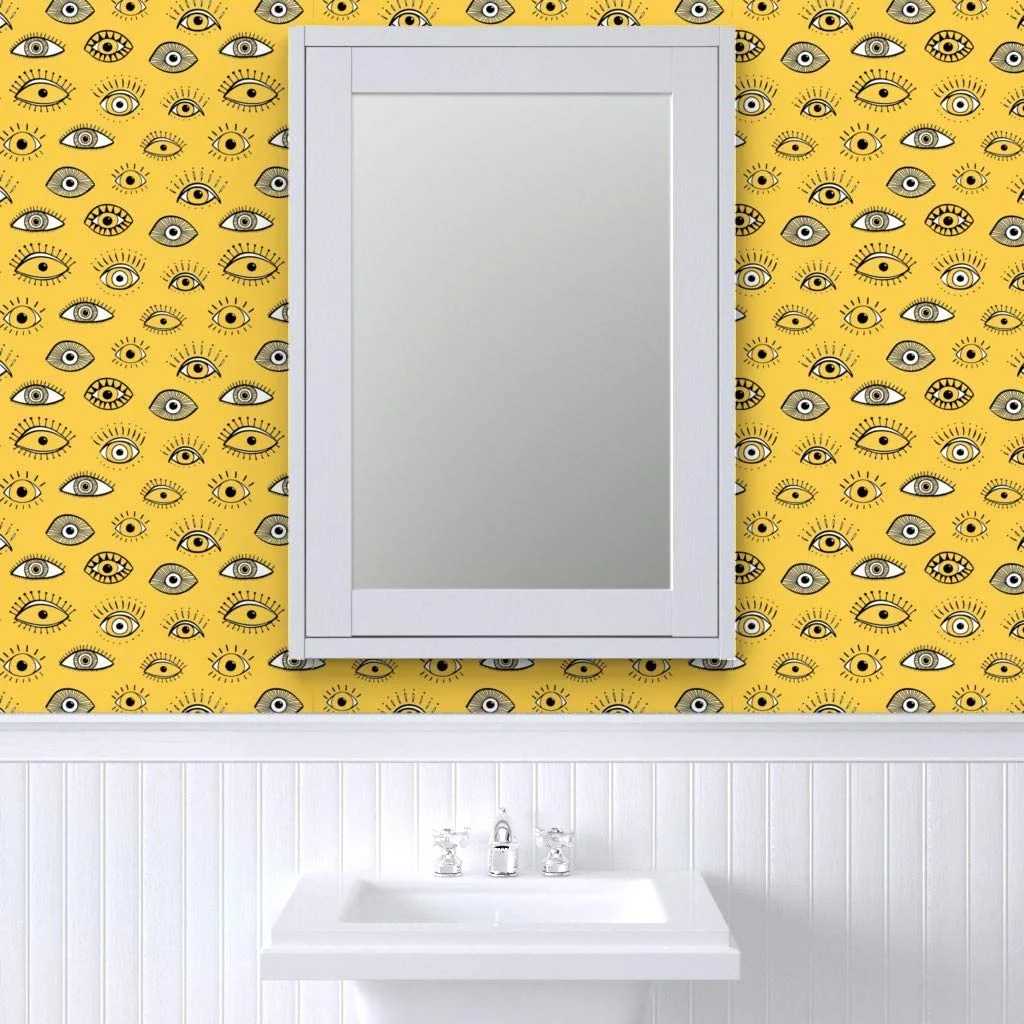 A white framed mirror and sink are set against a yellow wall featuring a black and white eye pattern above beadboard wainscoting.