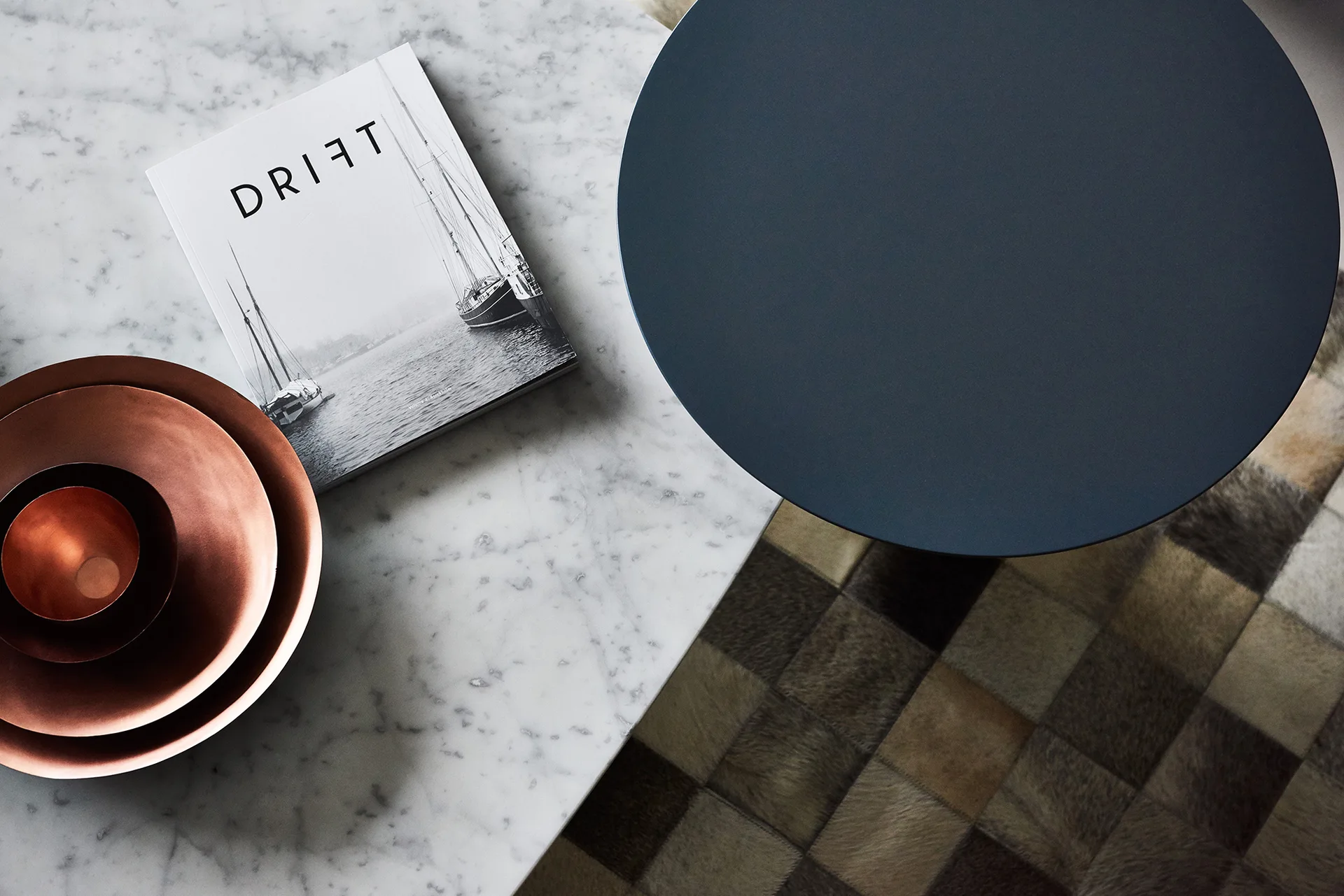 An overhead view of a white marble table with nested copper bowls and a book next to a matte navy circular side table on a checkered rug.