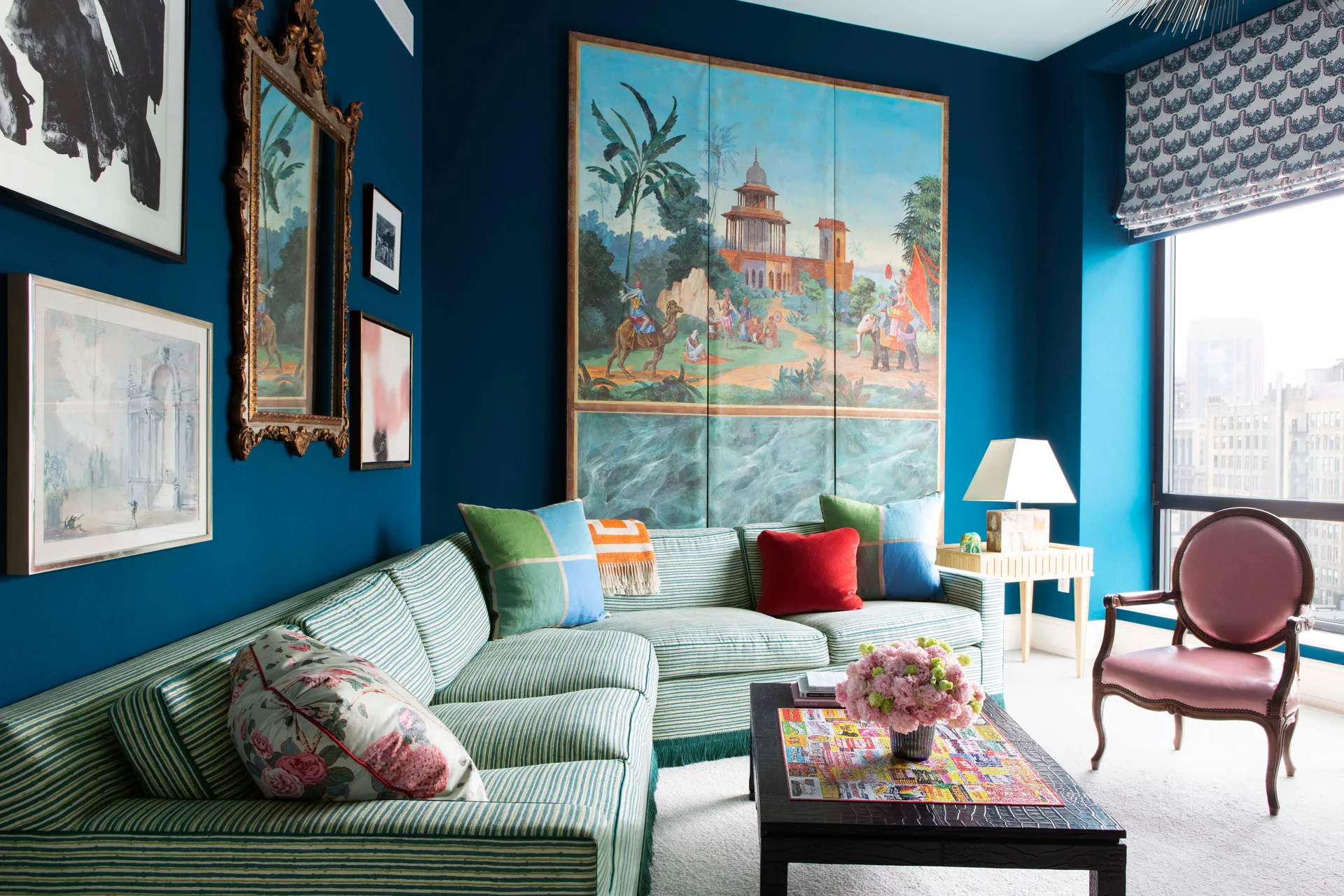 A green striped sectional sofa sits against a deep blue wall decorated with a large scenic painting, framed art prints, and an ornate gold mirror.