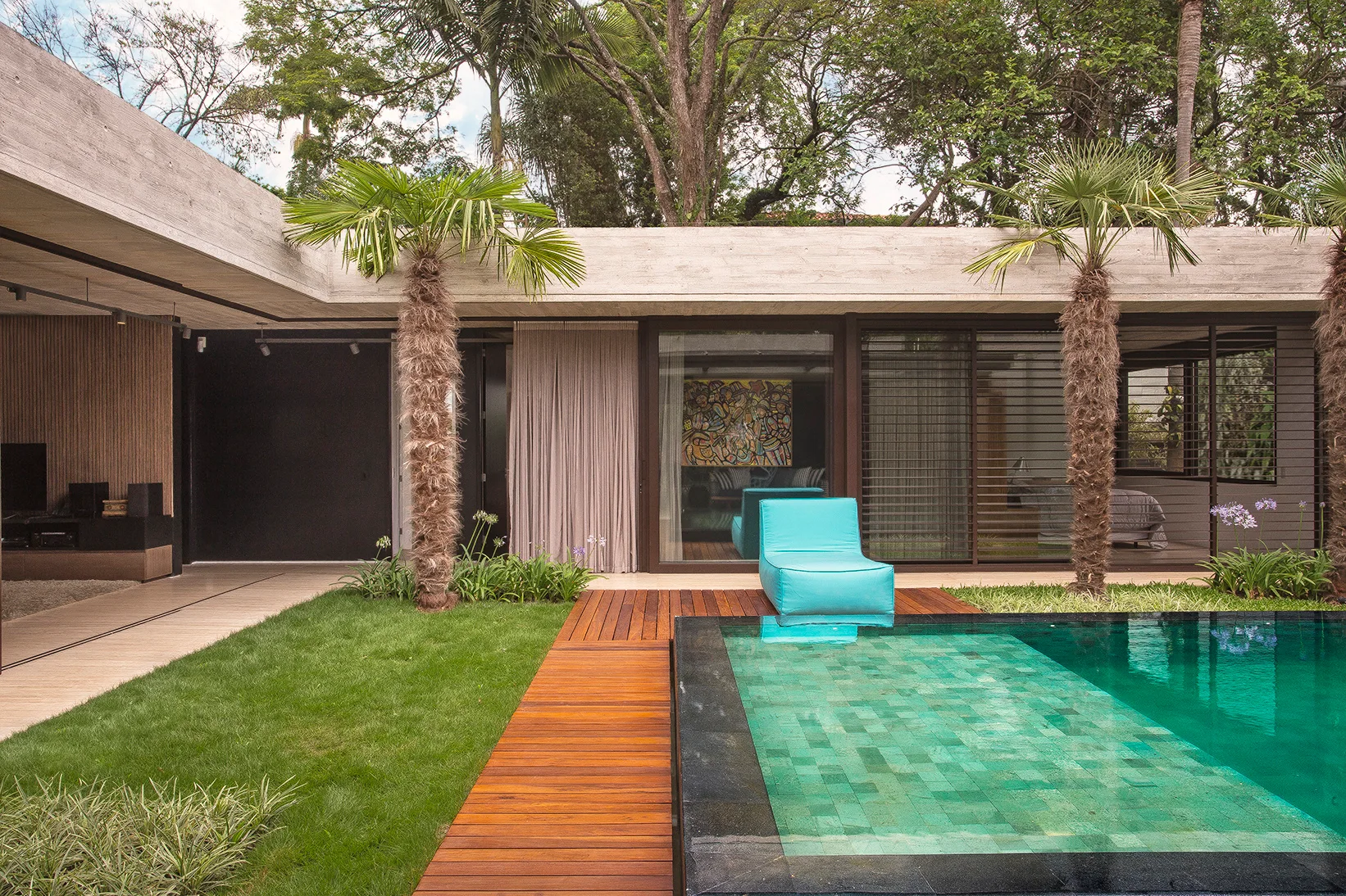 A modern concrete patio features a tiled swimming pool, a warm wood deck with a cyan lounge chair, a green lawn, and palm trees.