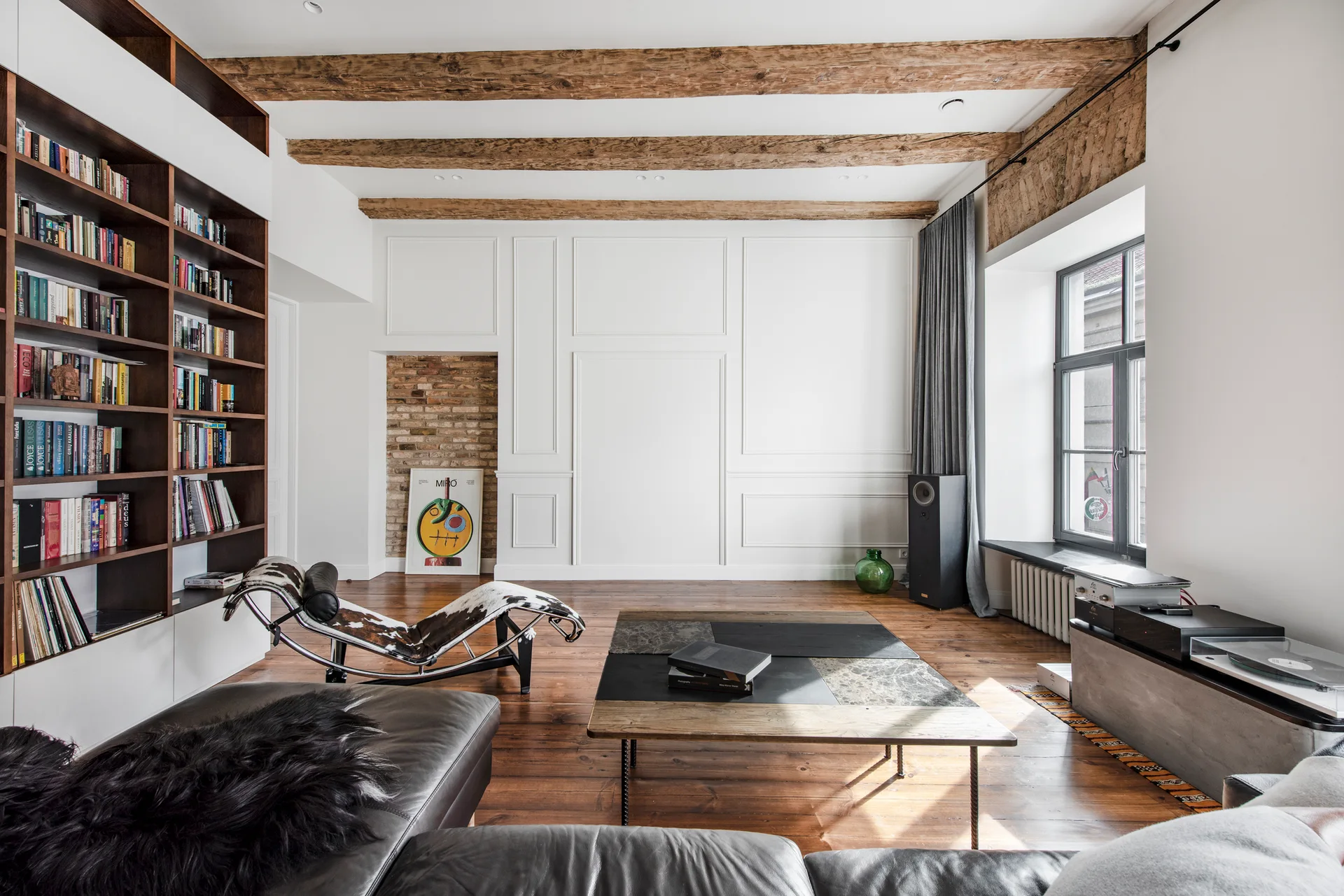 A living room features exposed wood ceiling beams, a tall dark wood bookshelf, a cowhide chaise lounge, and a leather sofa on wide plank wood floors.