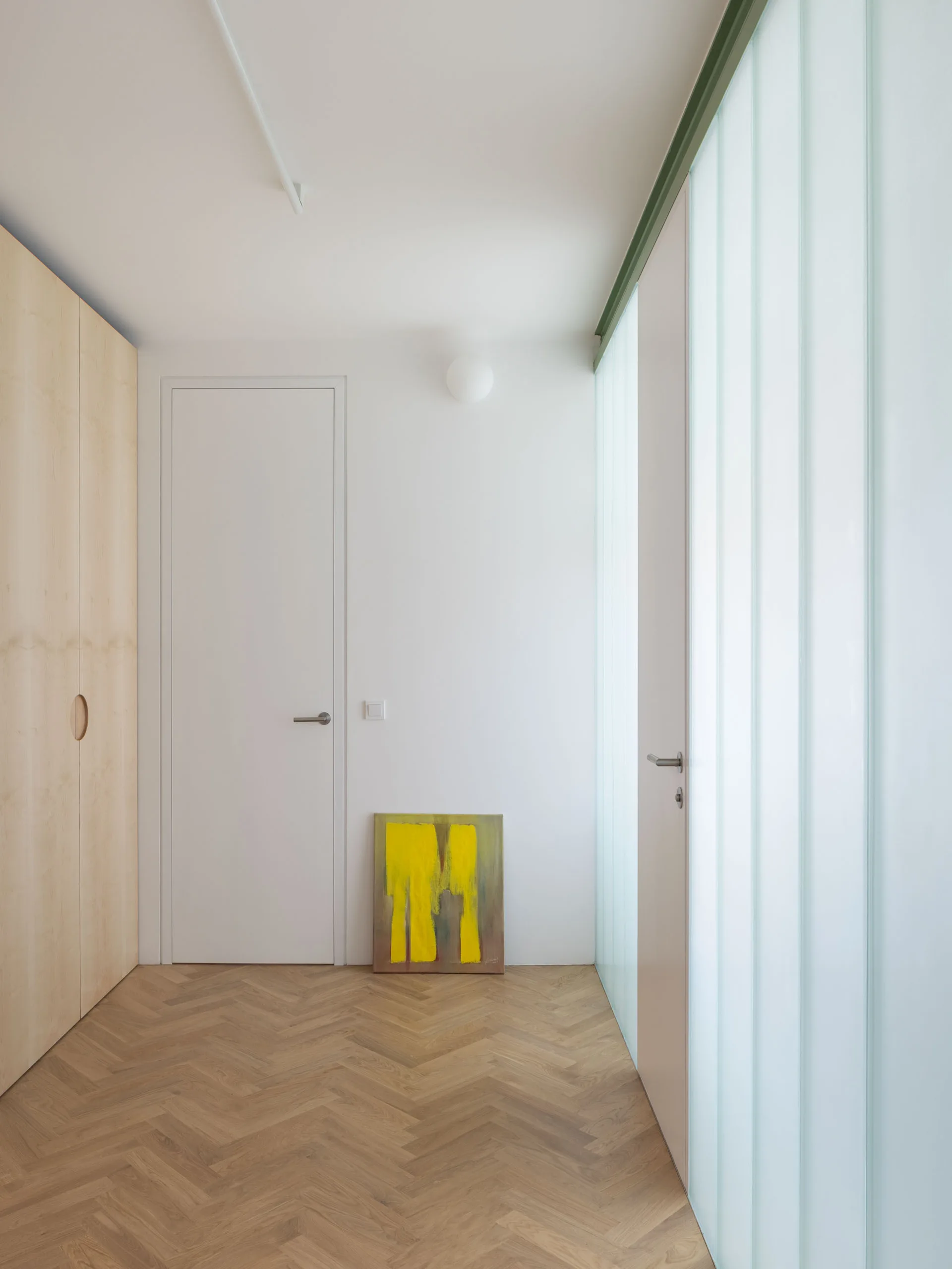 A minimalist hallway with light herringbone wood floors, a built-in wooden closet, a white door, and a translucent fluted glass wall.