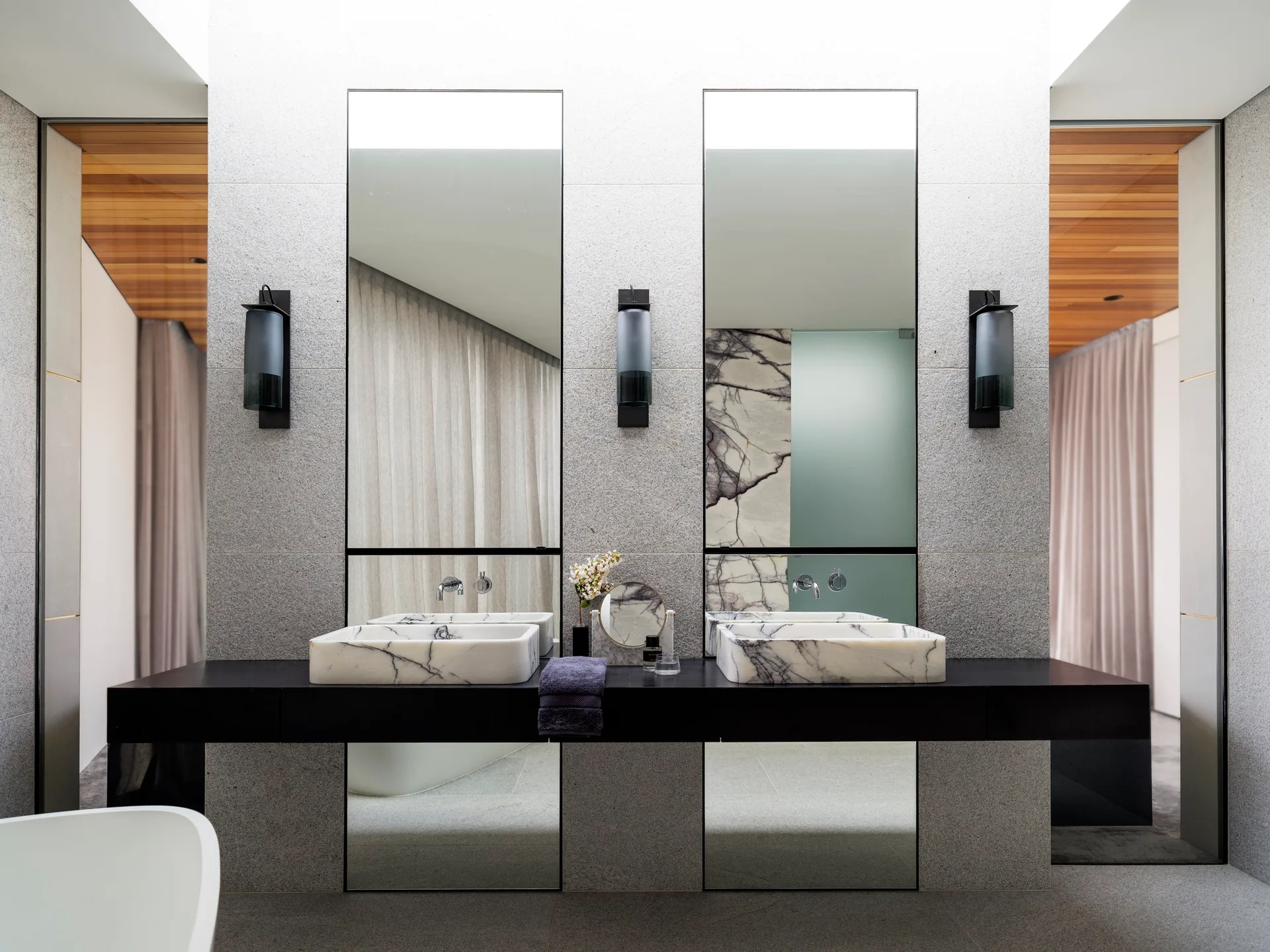 Two veined marble vessel sinks sit on a long black floating vanity beneath tall vertical mirrors and dark cylinder sconces on a grey stone wall.
