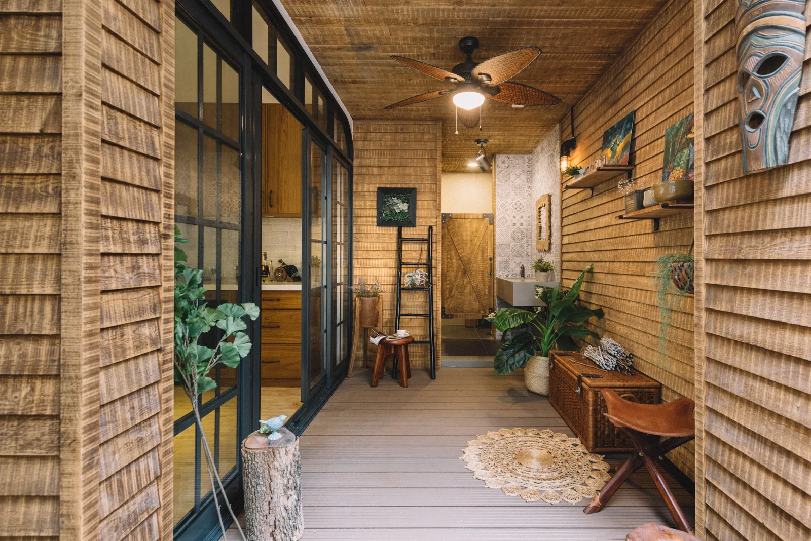 A rustic covered porch features horizontal wood siding, a plank ceiling with a fan, a wicker trunk, and black-framed glass sliding doors.