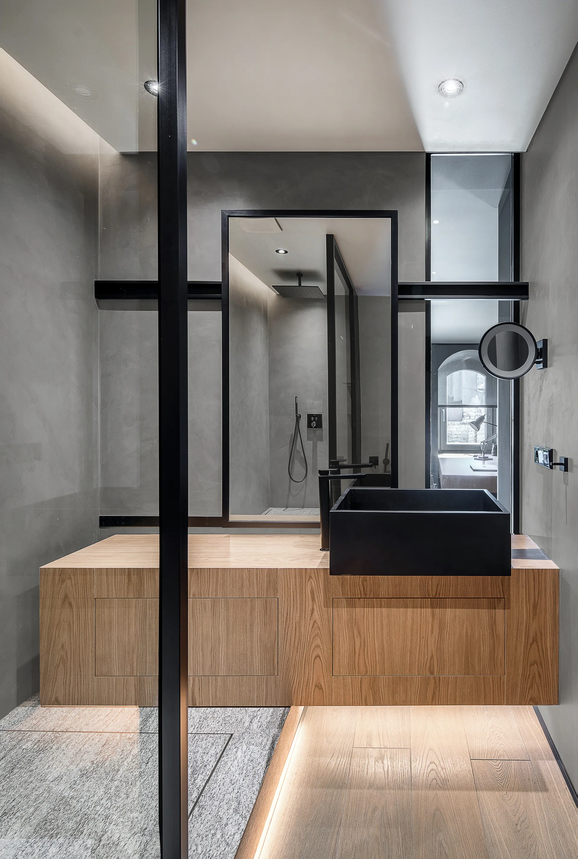 A modern bathroom features a wood vanity with a black vessel sink, concrete-finish walls, a large black-framed mirror, and a glass partition.