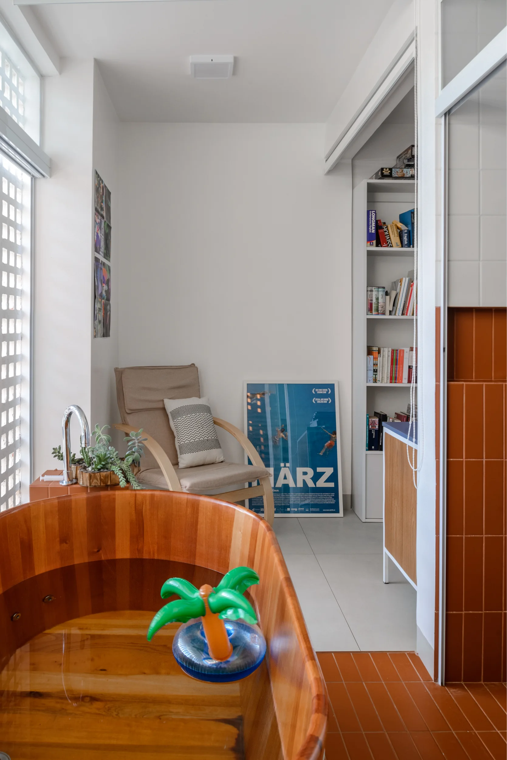 A contemporary bathroom features a wooden soaking tub, an armchair, a white bookshelf, and vertical terracotta wall tiles.