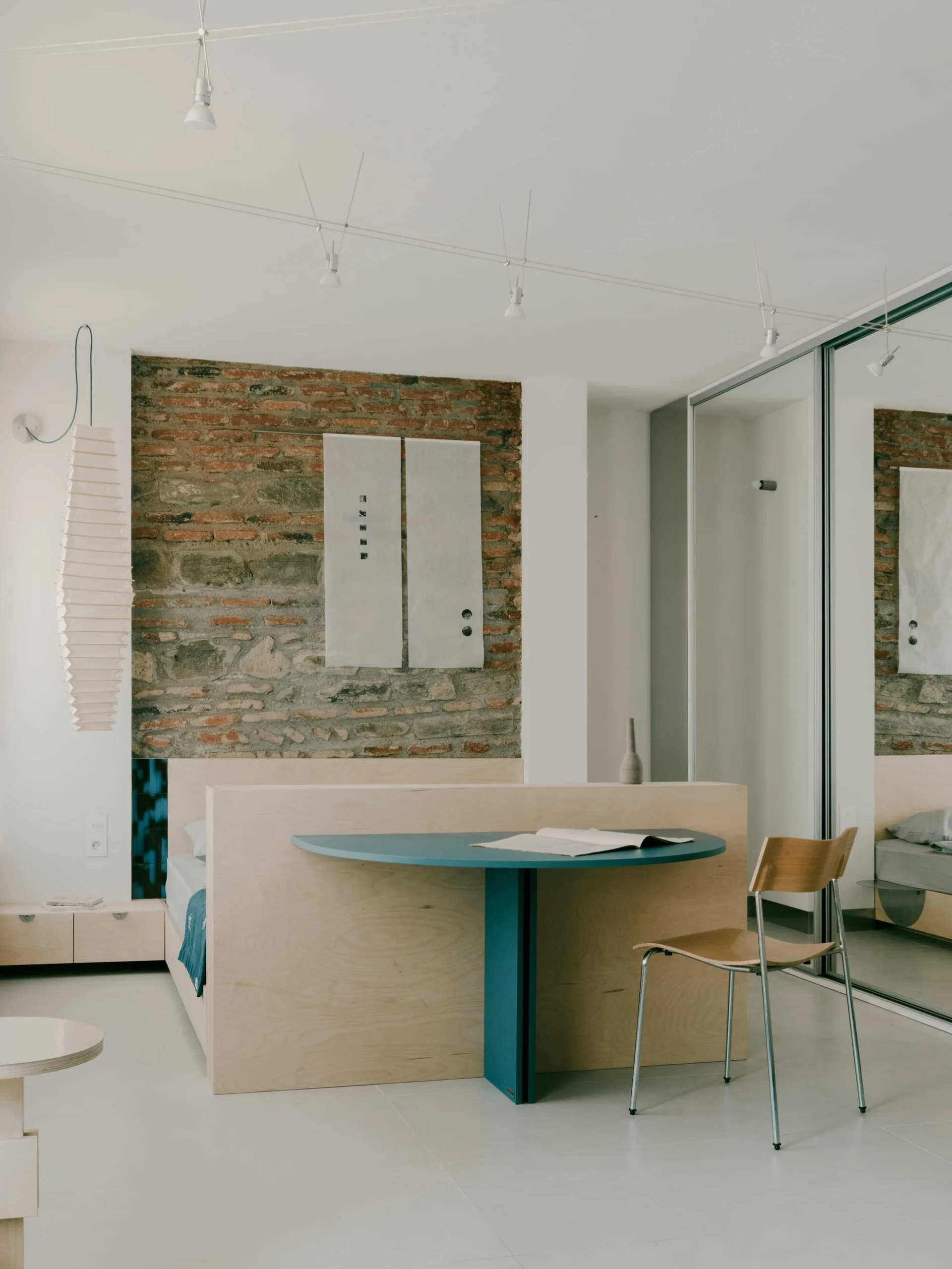 A minimalist bedroom features a custom plywood bed frame with an integrated teal desk, set against an exposed brick wall next to mirrored closet doors.
