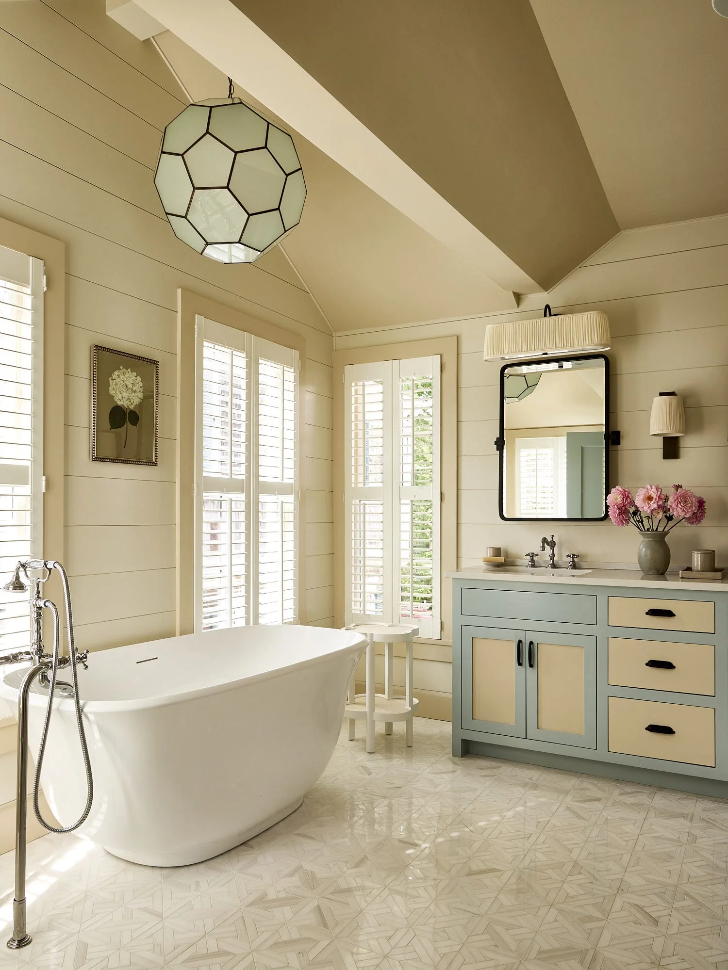 A cream bathroom featuring a white freestanding bathtub, a blue and cream vanity with a black-framed mirror, and patterned marble floor tile.