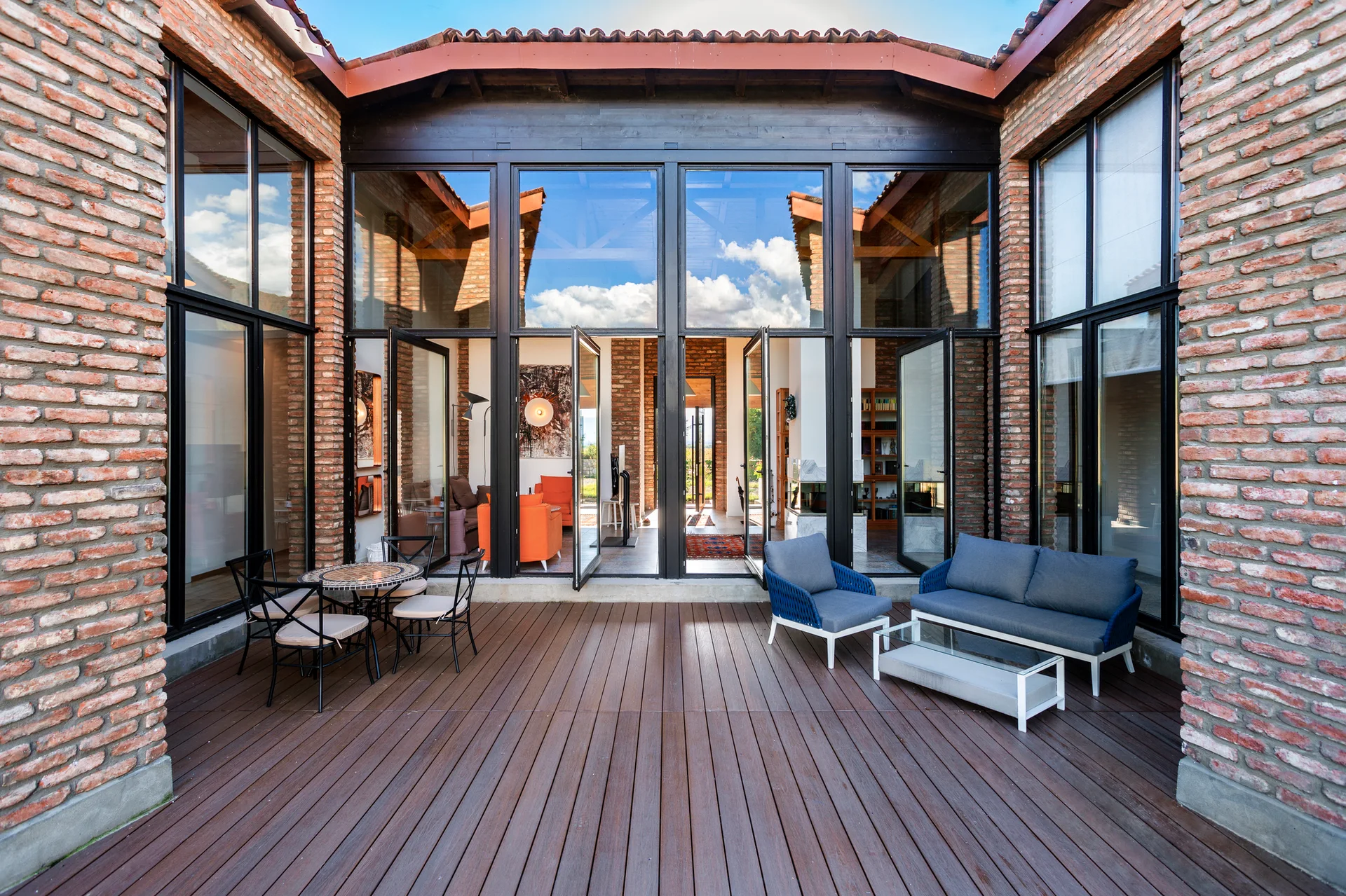 A wooden deck patio featuring a blue sofa and dining set is surrounded by exposed brick walls and large floor-to-ceiling glass doors.