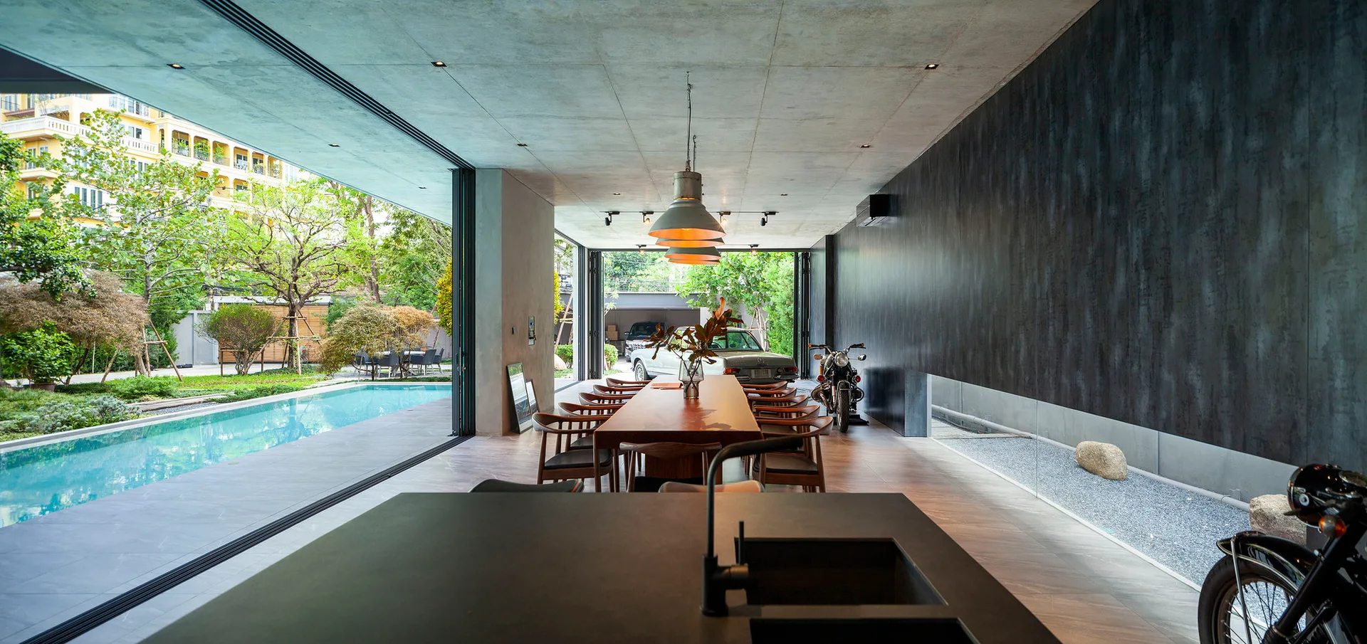 A long wooden dining table sits under industrial pendant lights in a concrete room with glass doors that open to an outdoor pool.