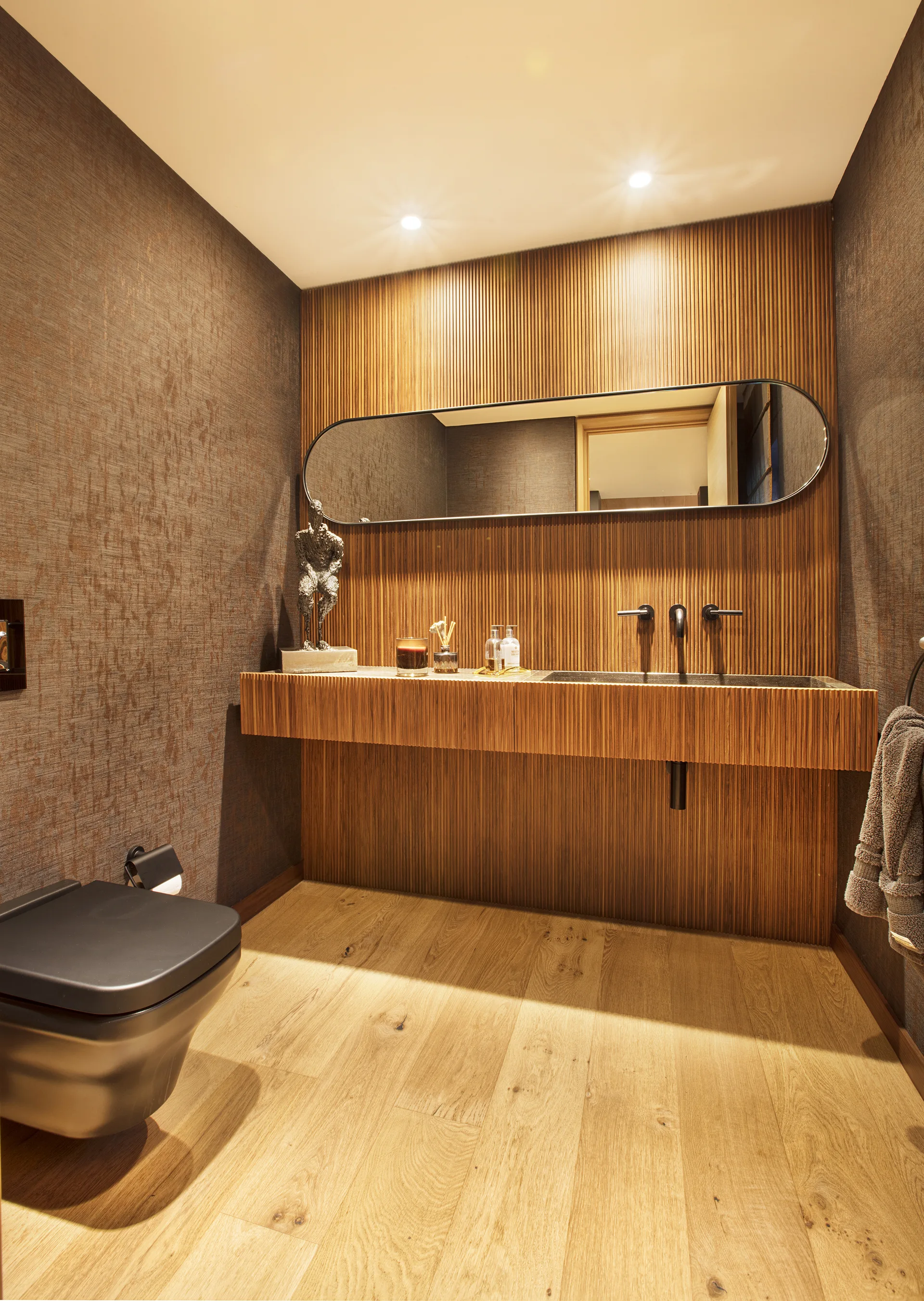 A modern powder room featuring a floating slatted wood vanity, a horizontal pill-shaped mirror, a black wall-mounted faucet, and textured wallpaper.