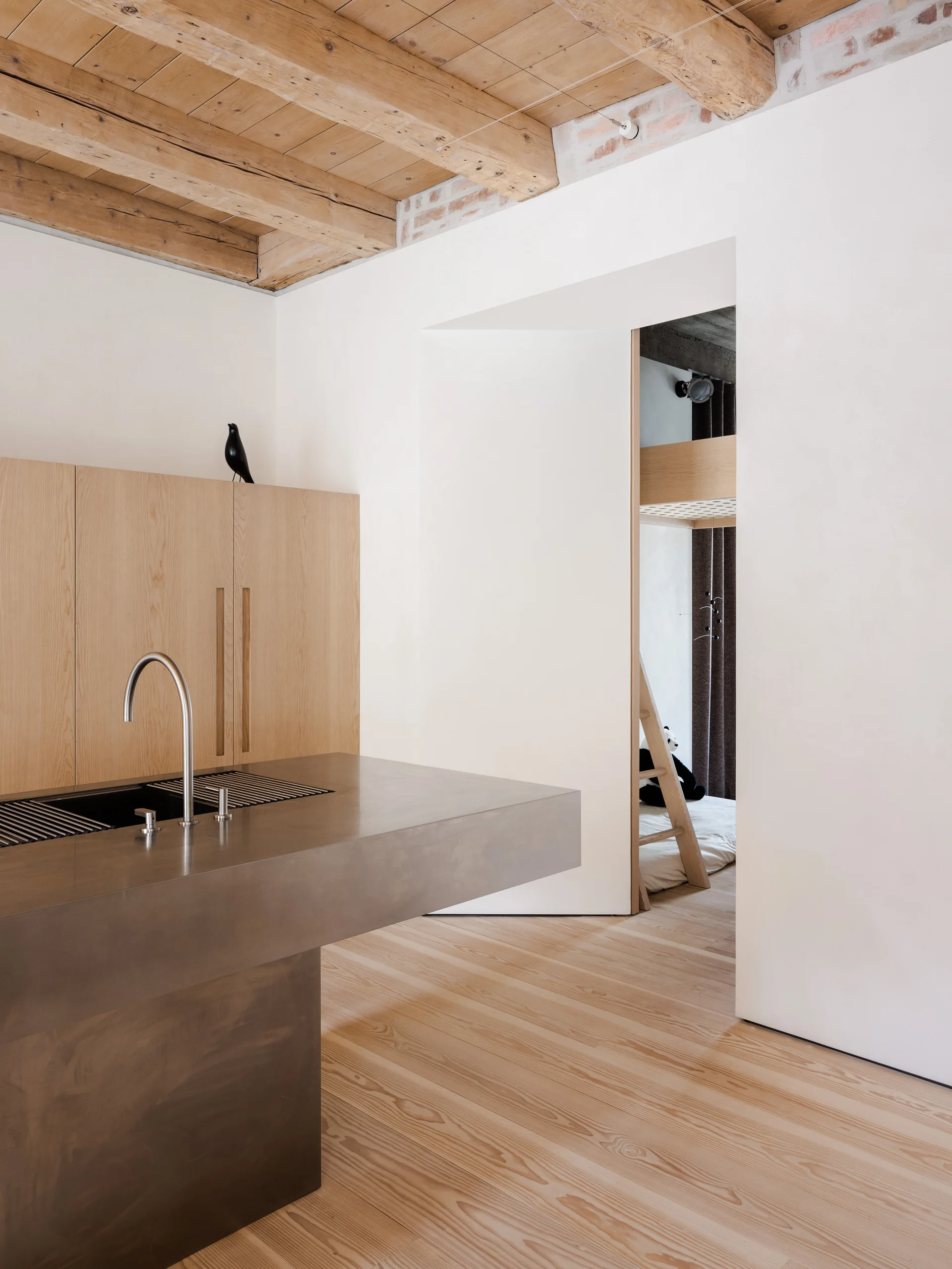 A minimalist kitchen features a matte metal island with an integrated sink, light wood cabinetry, white walls, and exposed wood ceiling beams.