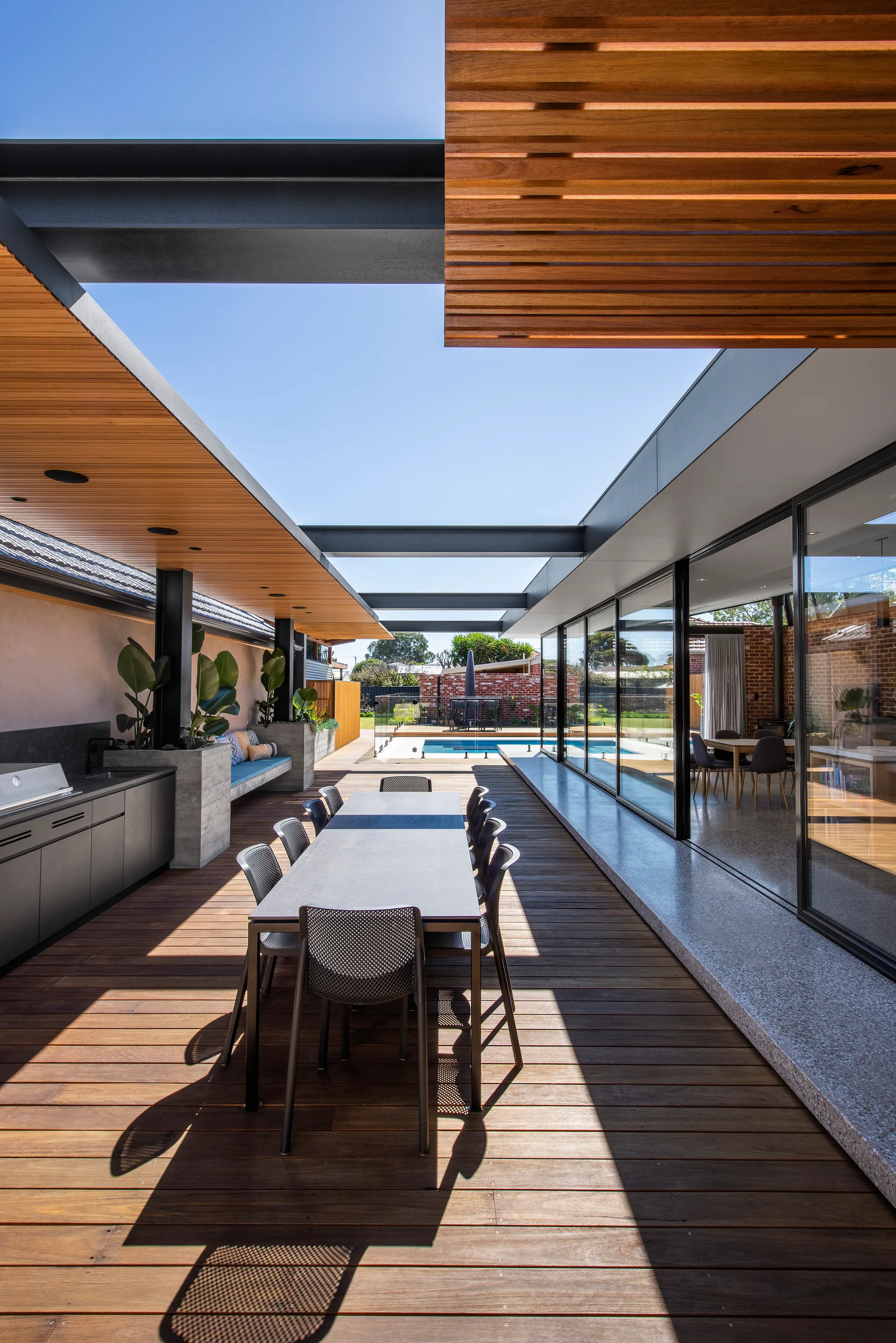 A modern wood patio deck features an outdoor dining table, built-in kitchen, slatted timber pergola, and a swimming pool visible in the background.