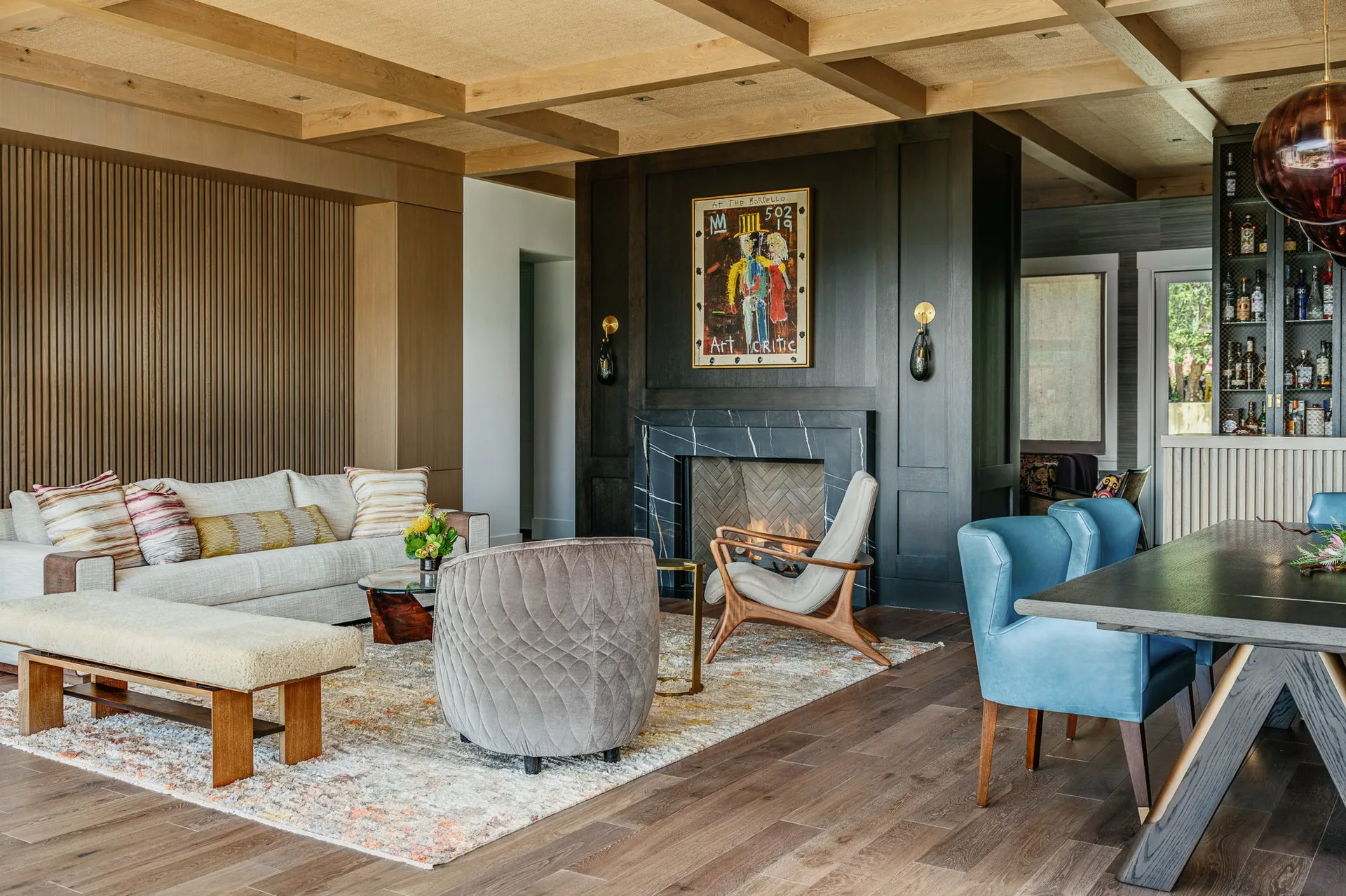 An open-concept living and dining area featuring a wood coffered ceiling, a dark paneled fireplace, a cream sofa, and blue dining chairs.