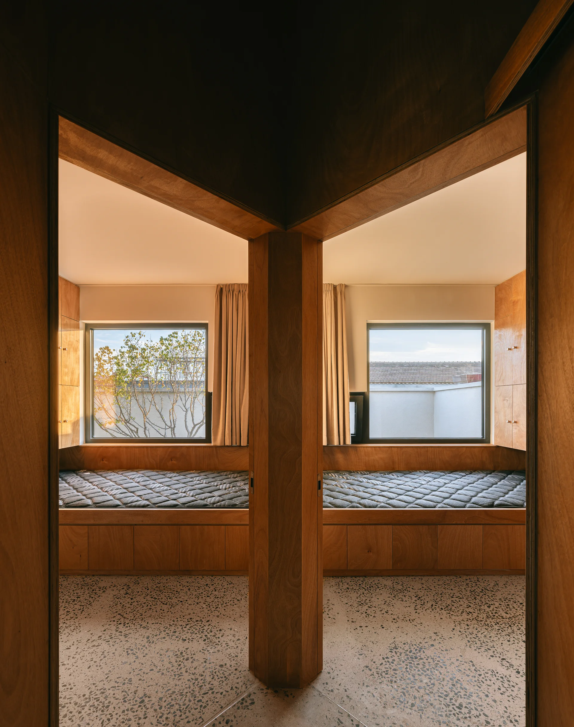 Two symmetrical wooden sleeping nooks with built-in beds are separated by a central column above speckled terrazzo flooring.