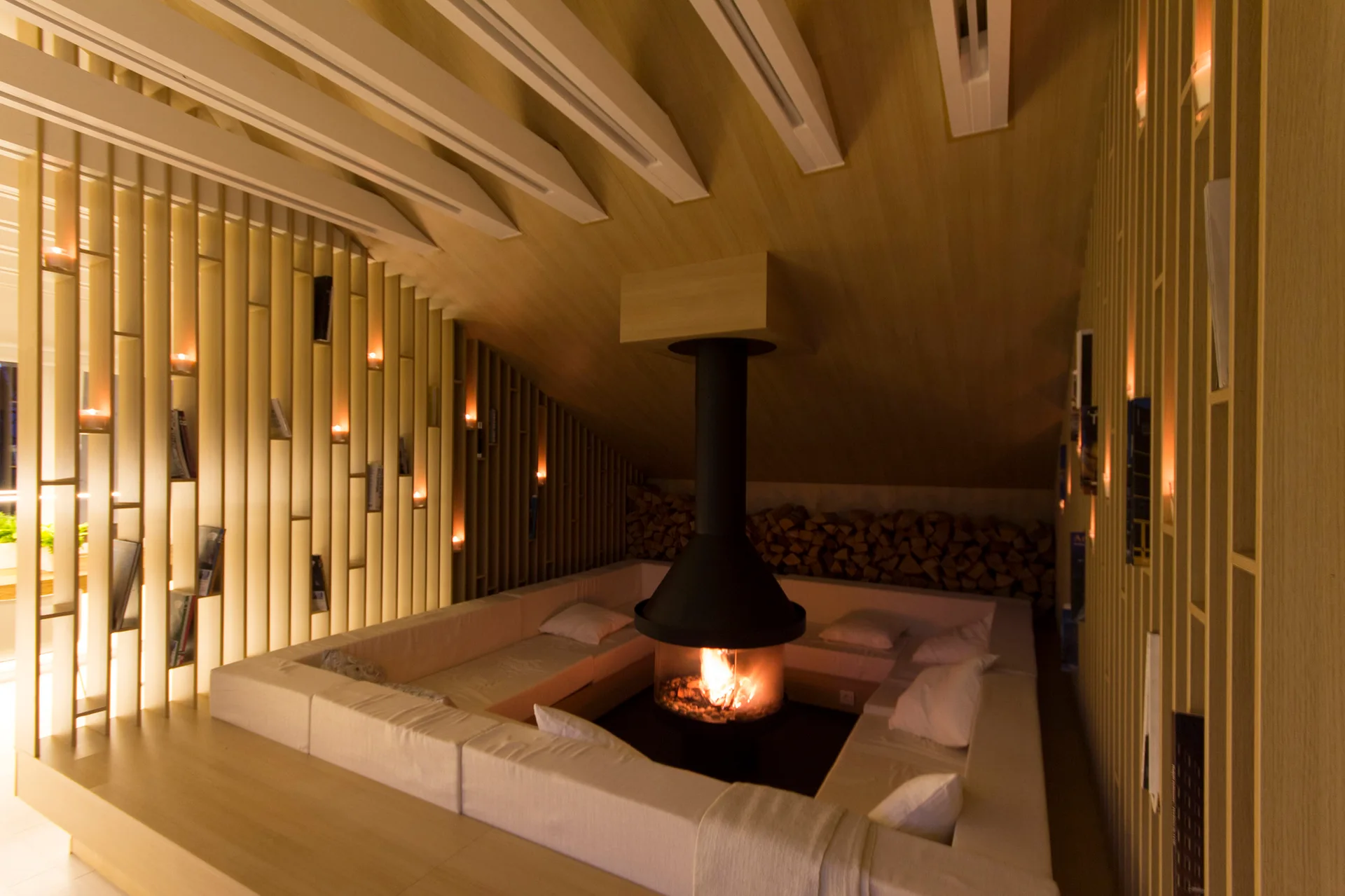 A sunken conversation pit with white built-in seating surrounds a central black hanging fireplace in a room with sloped wood paneling.