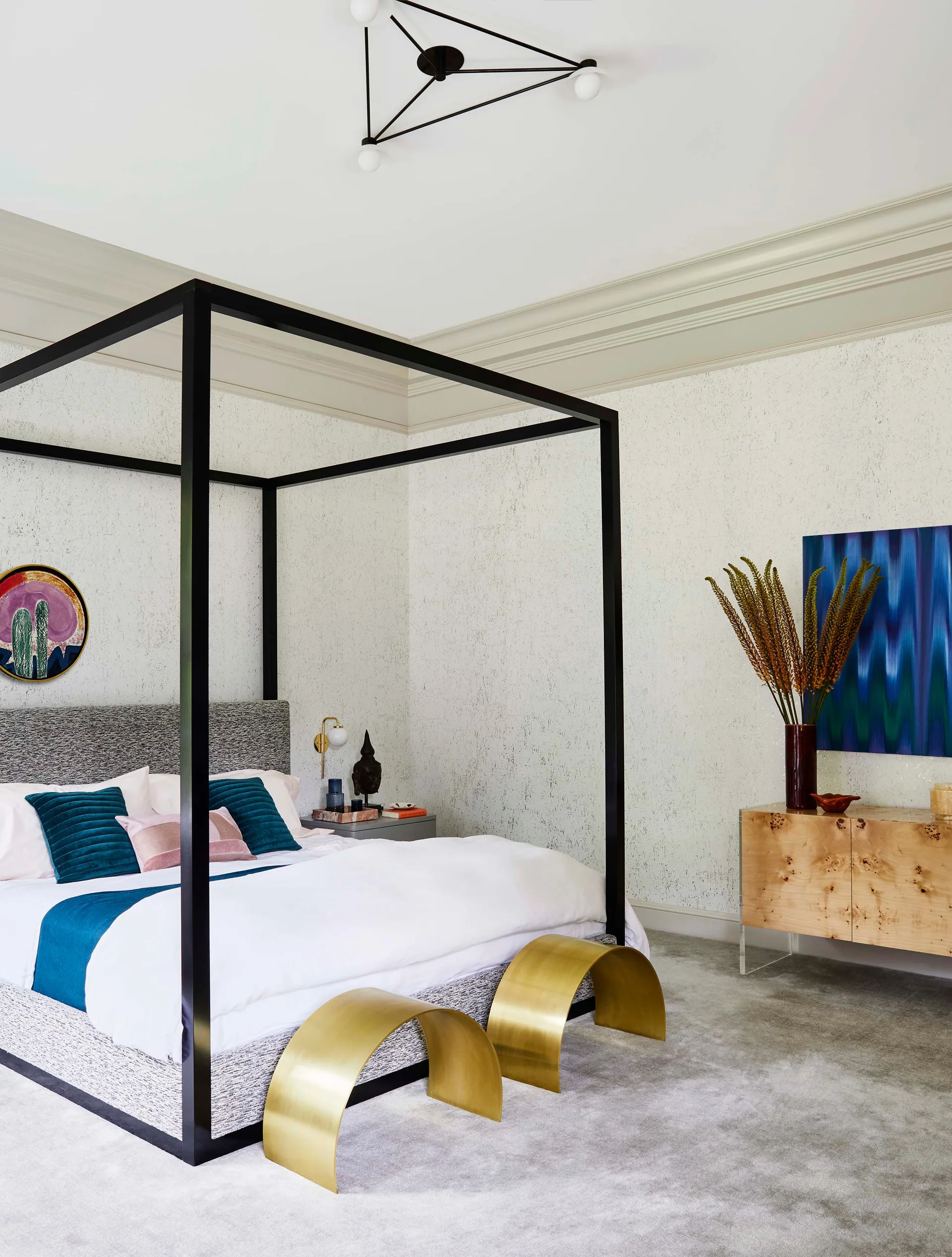 A modern bedroom features a black canopy bed with teal pillows, flanked by curved brass stools and a burl wood dresser.