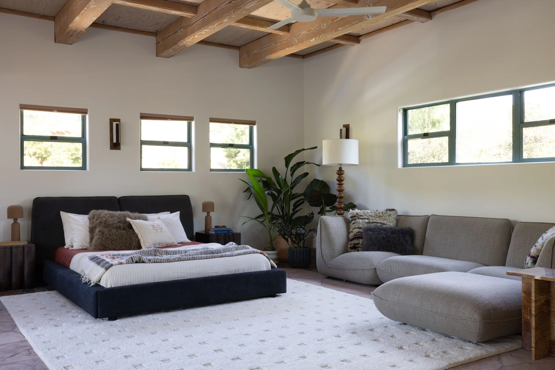 A spacious bedroom features an upholstered dark bed, a low-slung modular sofa, a cream shag rug, and exposed wood ceiling beams.