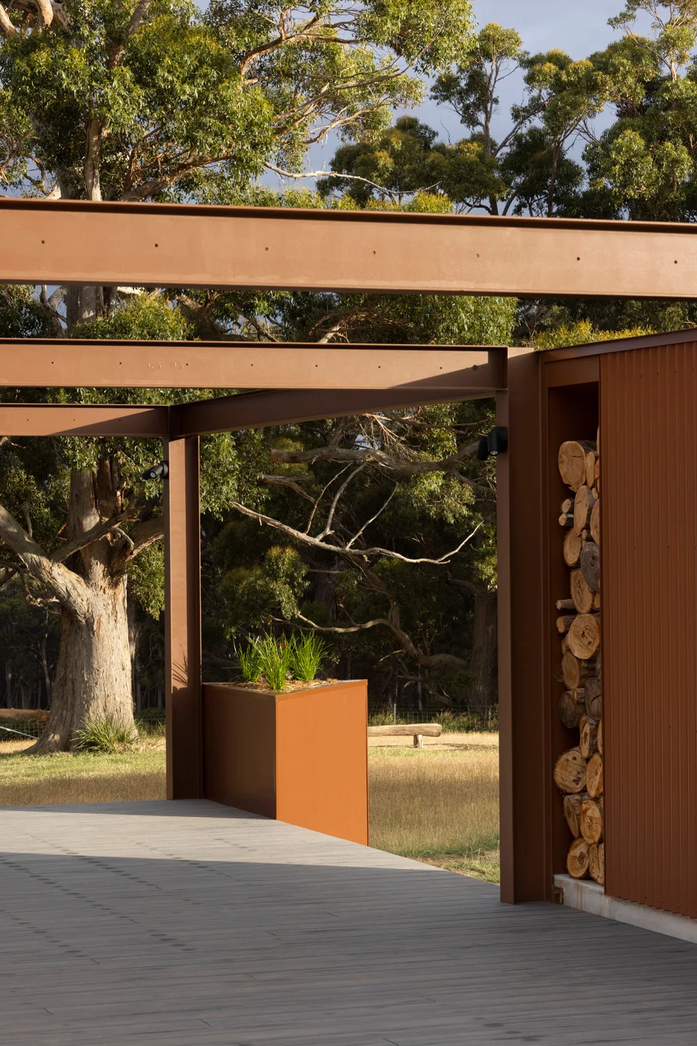 An outdoor patio featuring rust-colored steel beams, a gray plank deck, a metal planter, and a built-in niche filled with stacked firewood.
