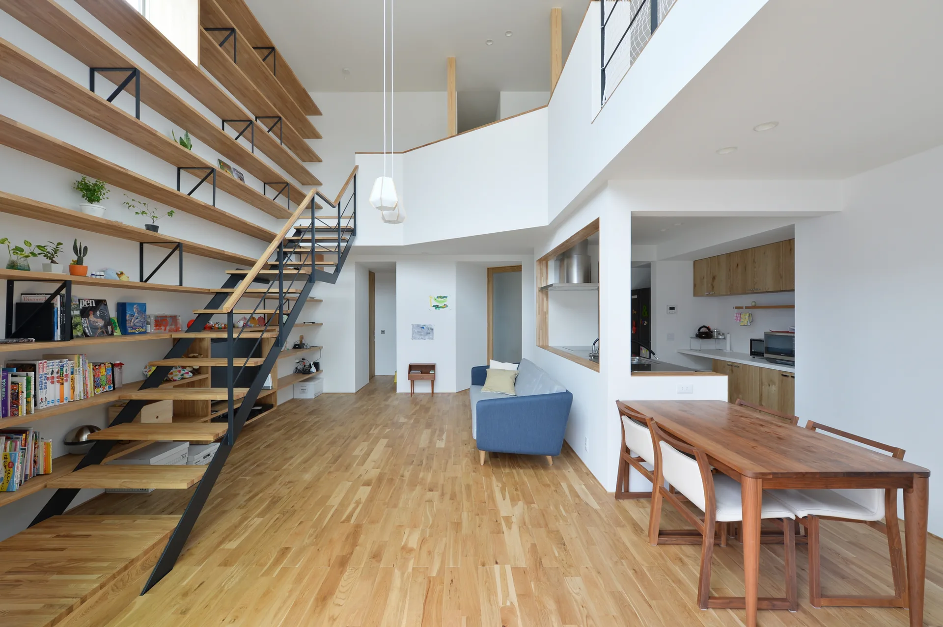 An open-plan residential interior features a large wooden bookshelf wall with an integrated black metal staircase, a blue sofa, and a wooden dining table.