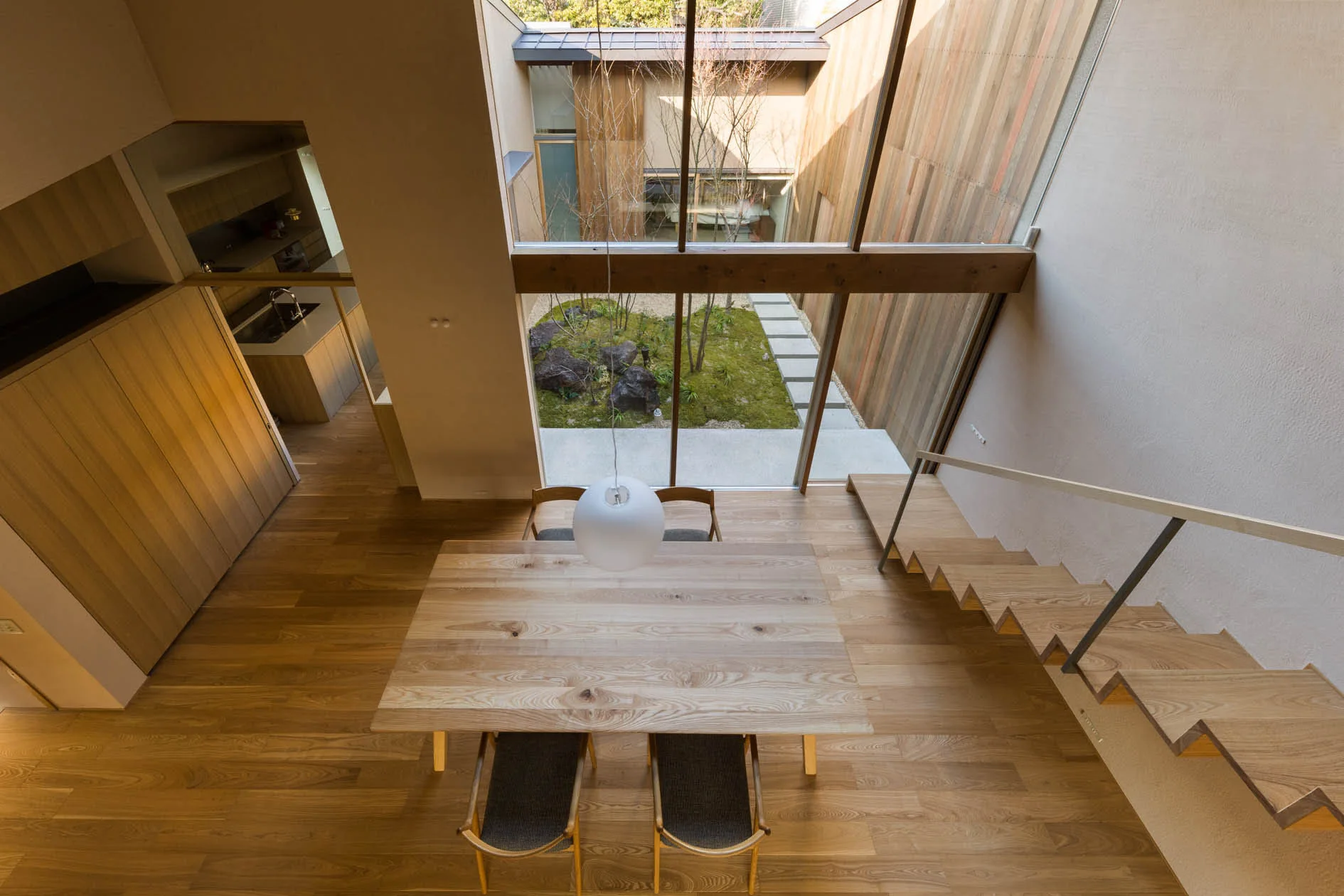 A light wood dining table with chairs sits next to a floating wooden staircase facing a large glass window that overlooks a green internal courtyard.