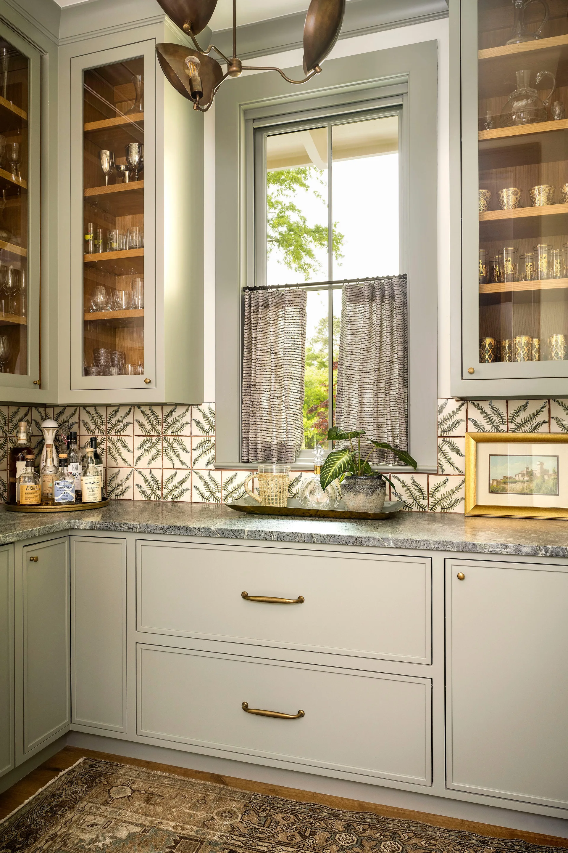 Sage green cabinets with glass fronts flank a central window above a stone countertop and fern-patterned tile backsplash.