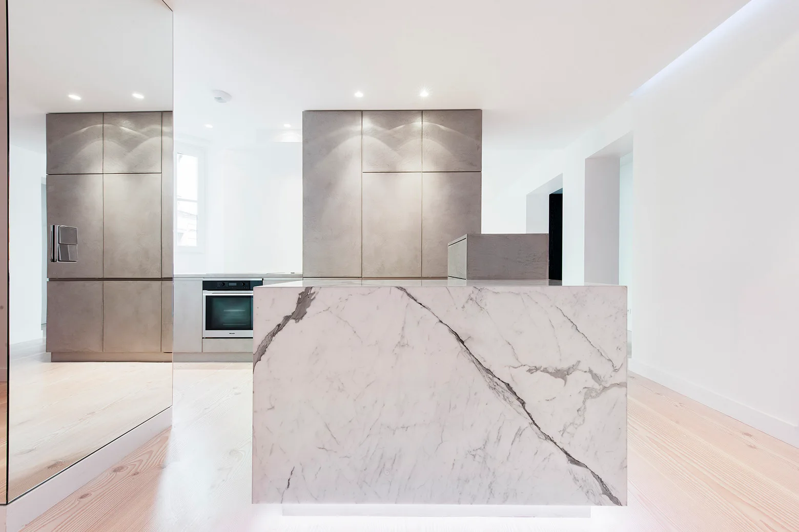 A minimalist modern kitchen features a large white-veined marble island, seamless gray cabinetry, light wood floors, and a mirrored wall.