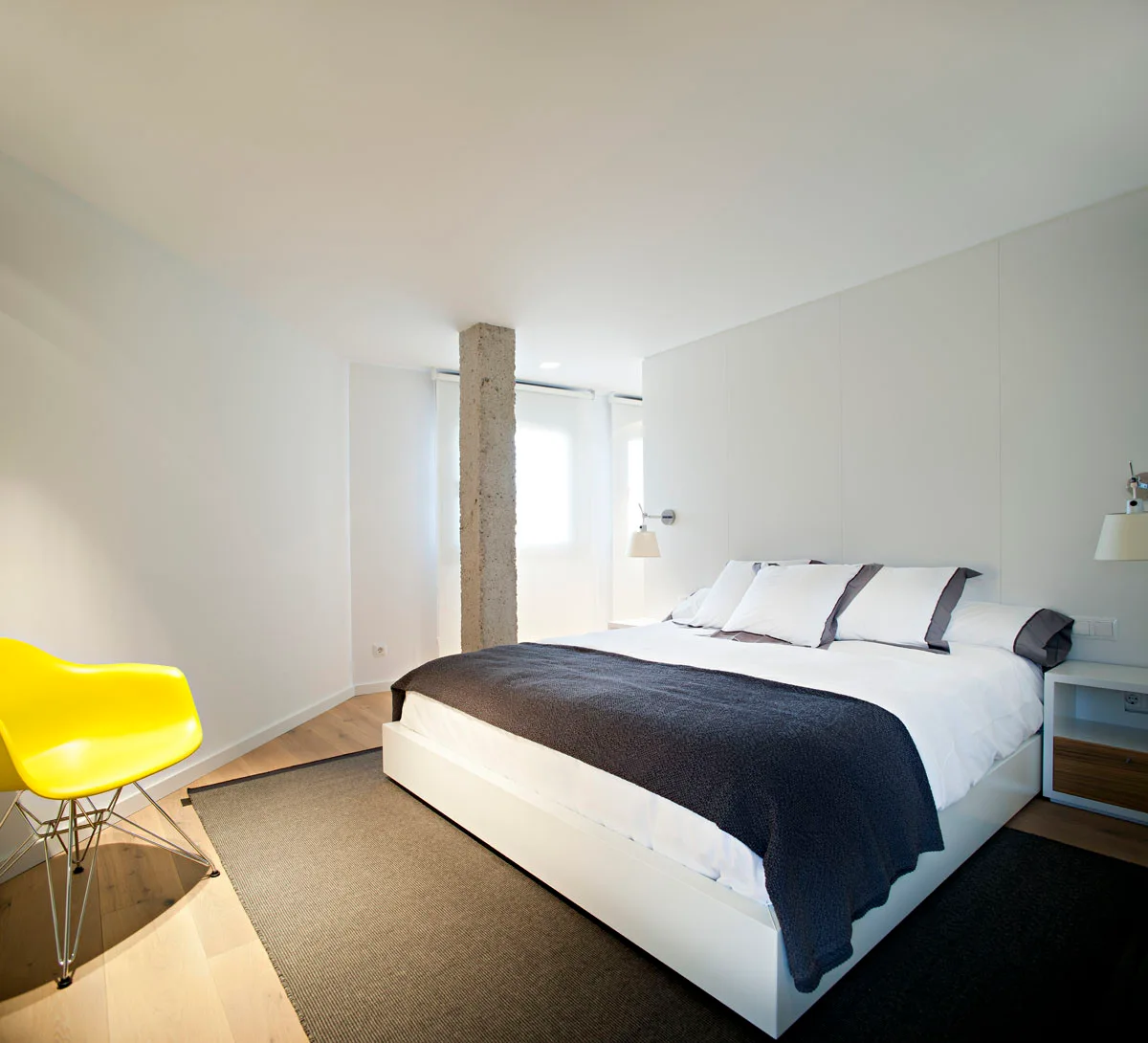 A minimalist bedroom featuring a white bed with grey accents, a textured concrete column, and a bright yellow molded plastic chair on wood floors.