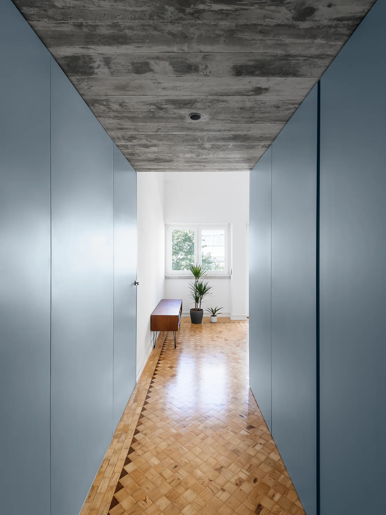 A narrow hallway with seamless blue wall-to-wall cabinets, a raw concrete ceiling, and a patterned wood parquet floor leading to a window.