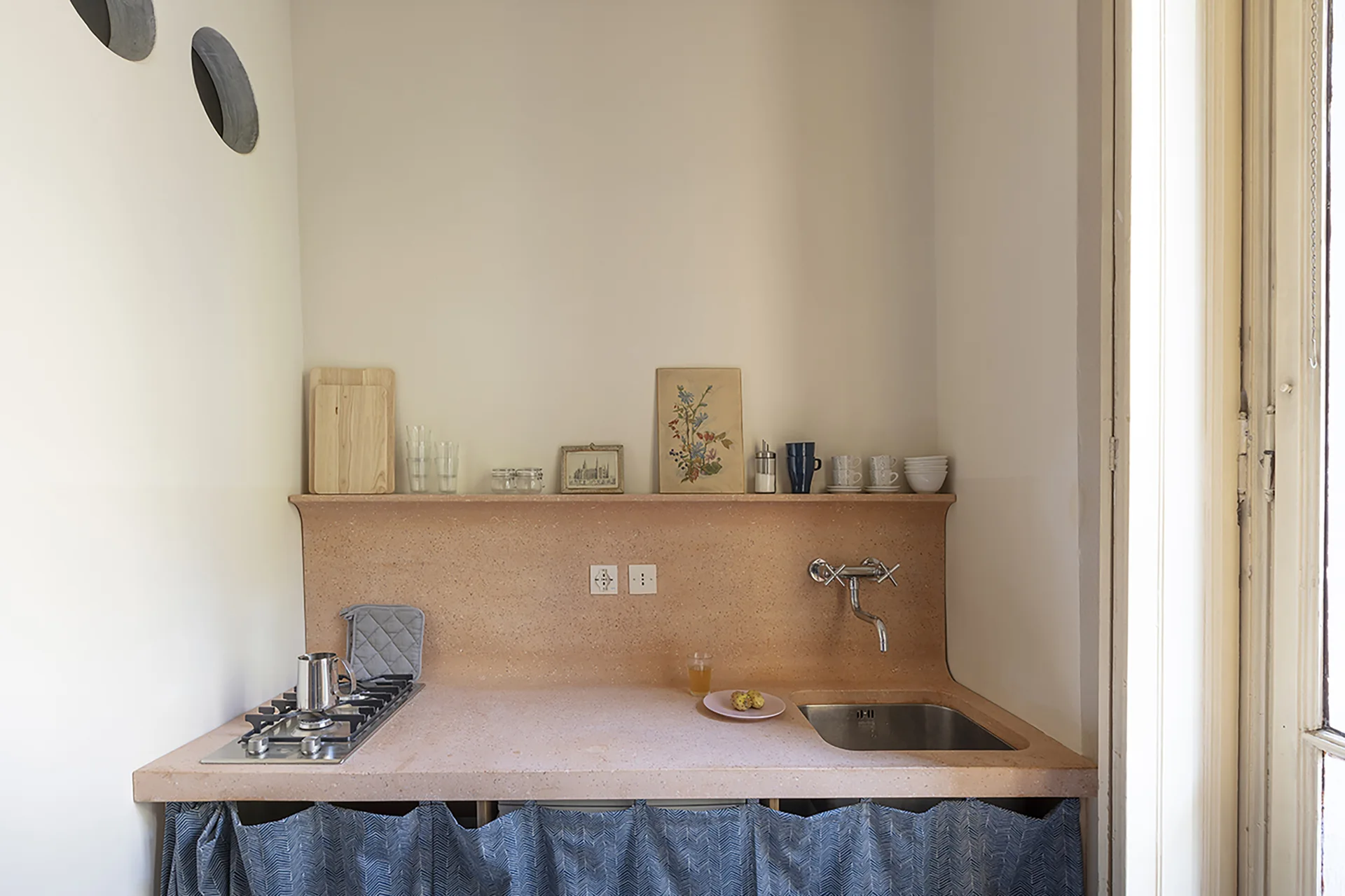 A small kitchen featuring a peach-colored composite countertop, a wall-mounted faucet, a metal gas hob, and a blue patterned curtain skirt below.