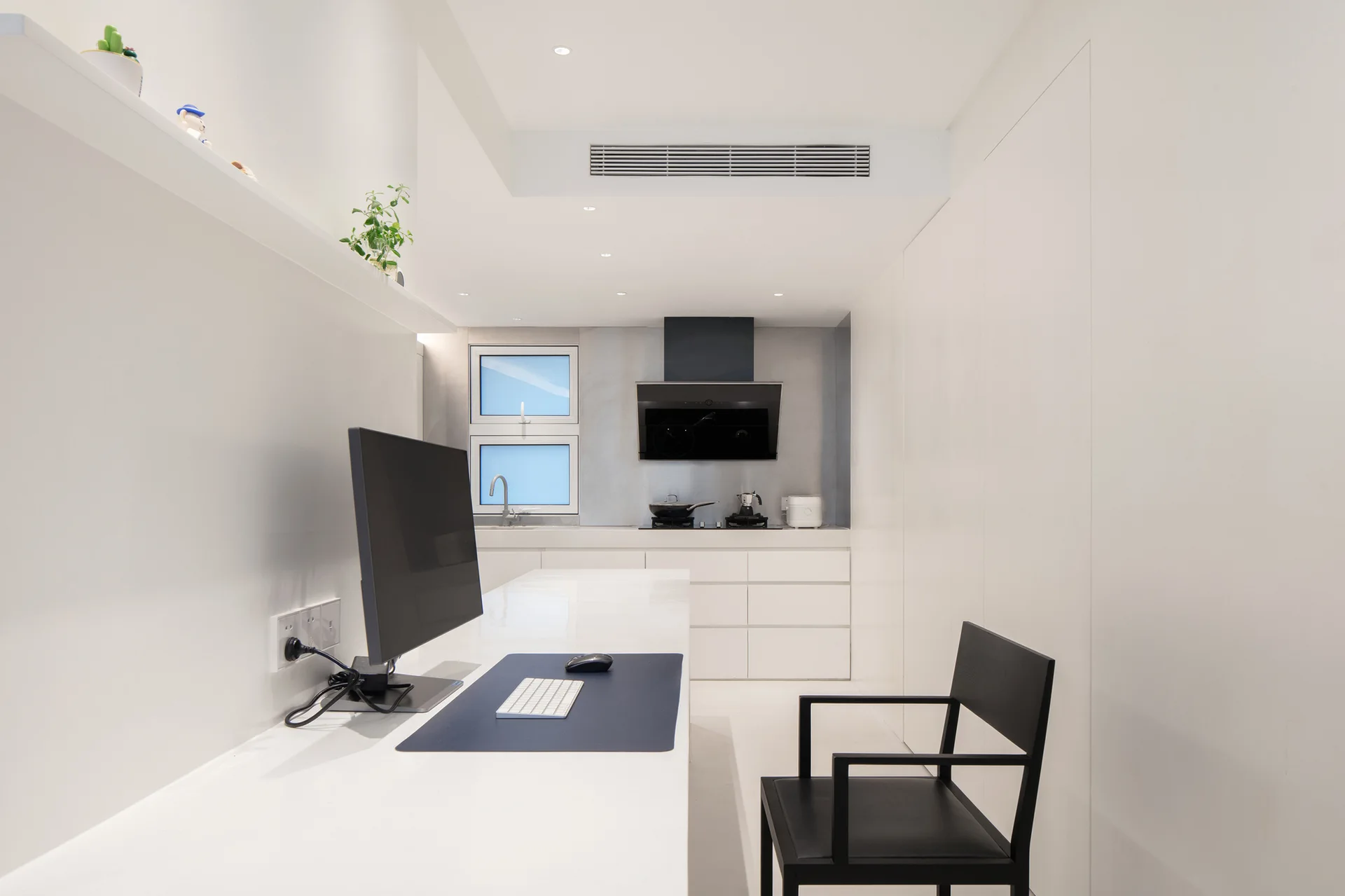 A long white desk with a computer monitor and black chair integrates into a minimalist white kitchen with flat-front cabinets and a black range hood.