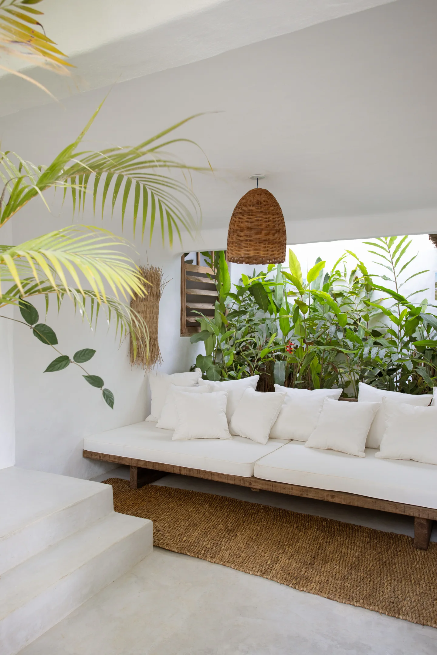 A low wooden daybed with white cushions sits on a woven rug beneath a hanging wicker pendant light in a tropical outdoor lounge.