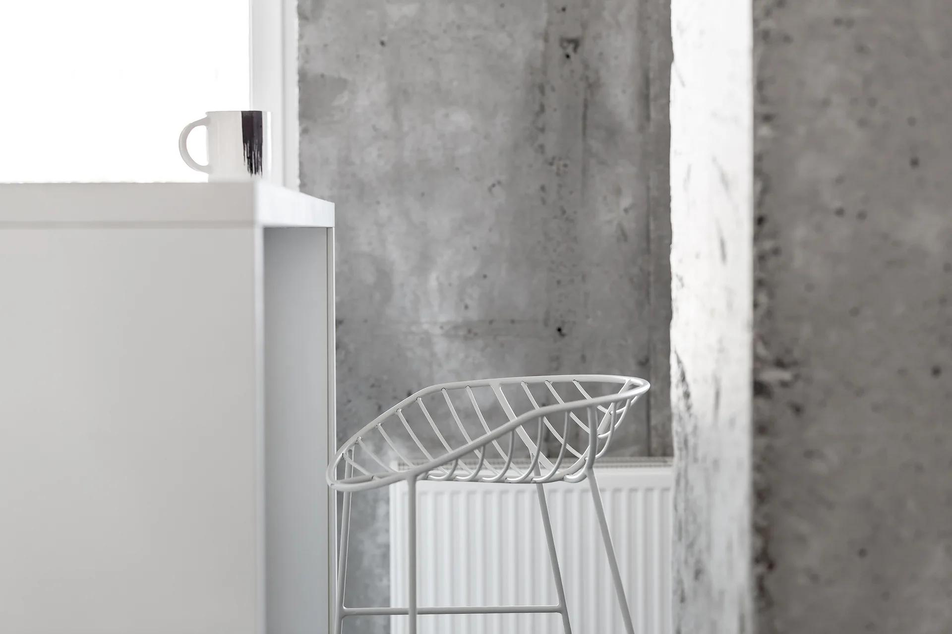 A white wire counter stool stands beside a white table against a rough grey concrete wall under bright natural light.